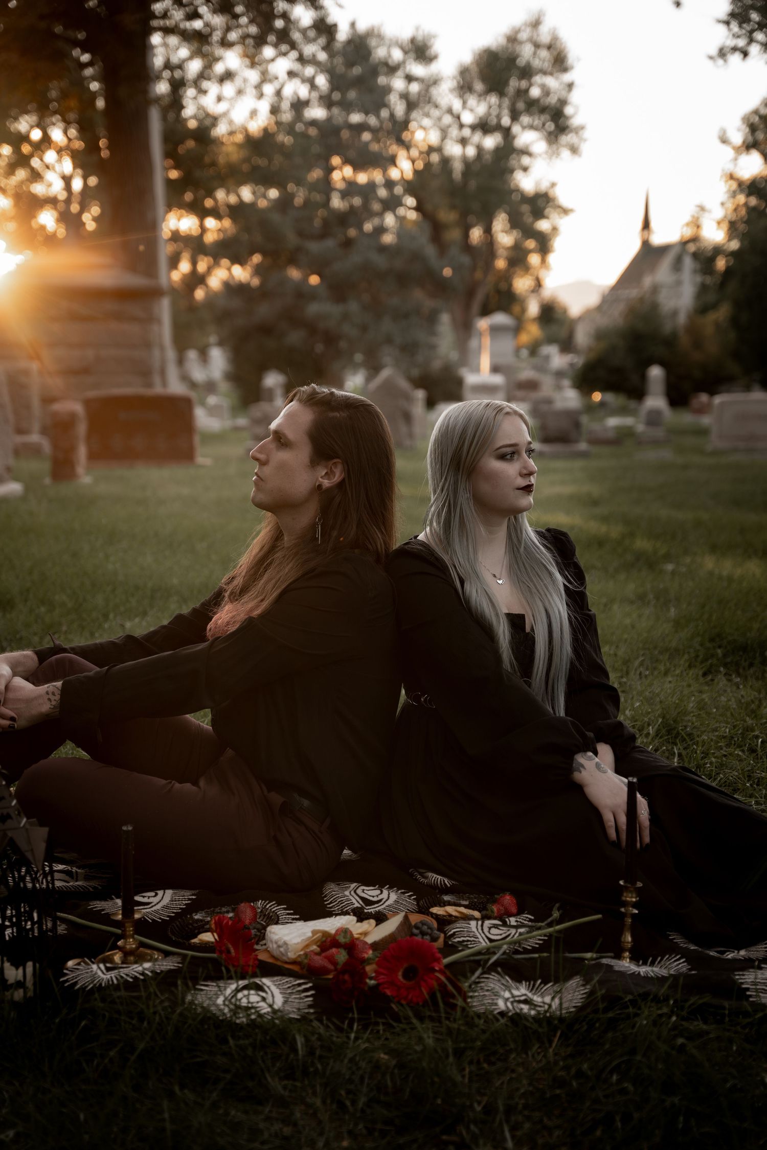 Two goths in black sit back-to-back on a blanket in a cemetery at sunset with red flowers nearby.