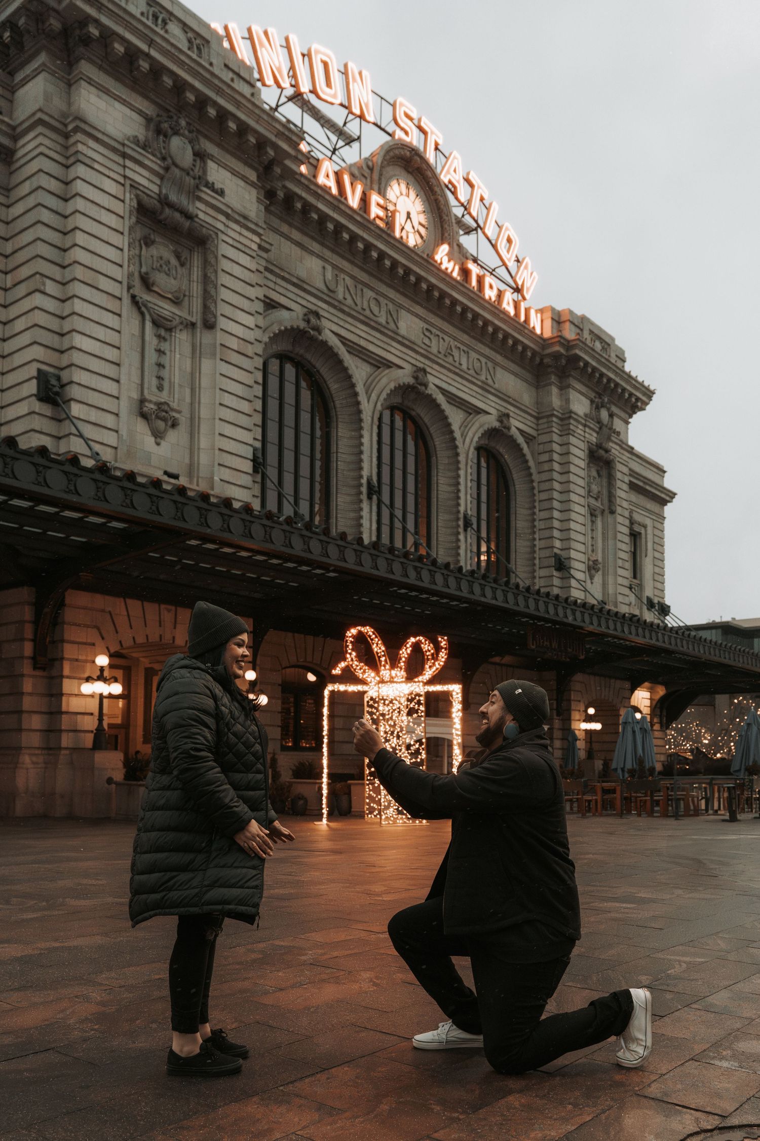 Man proposing on one knee to woman outside Union Station Denver with holiday lights at dusk.