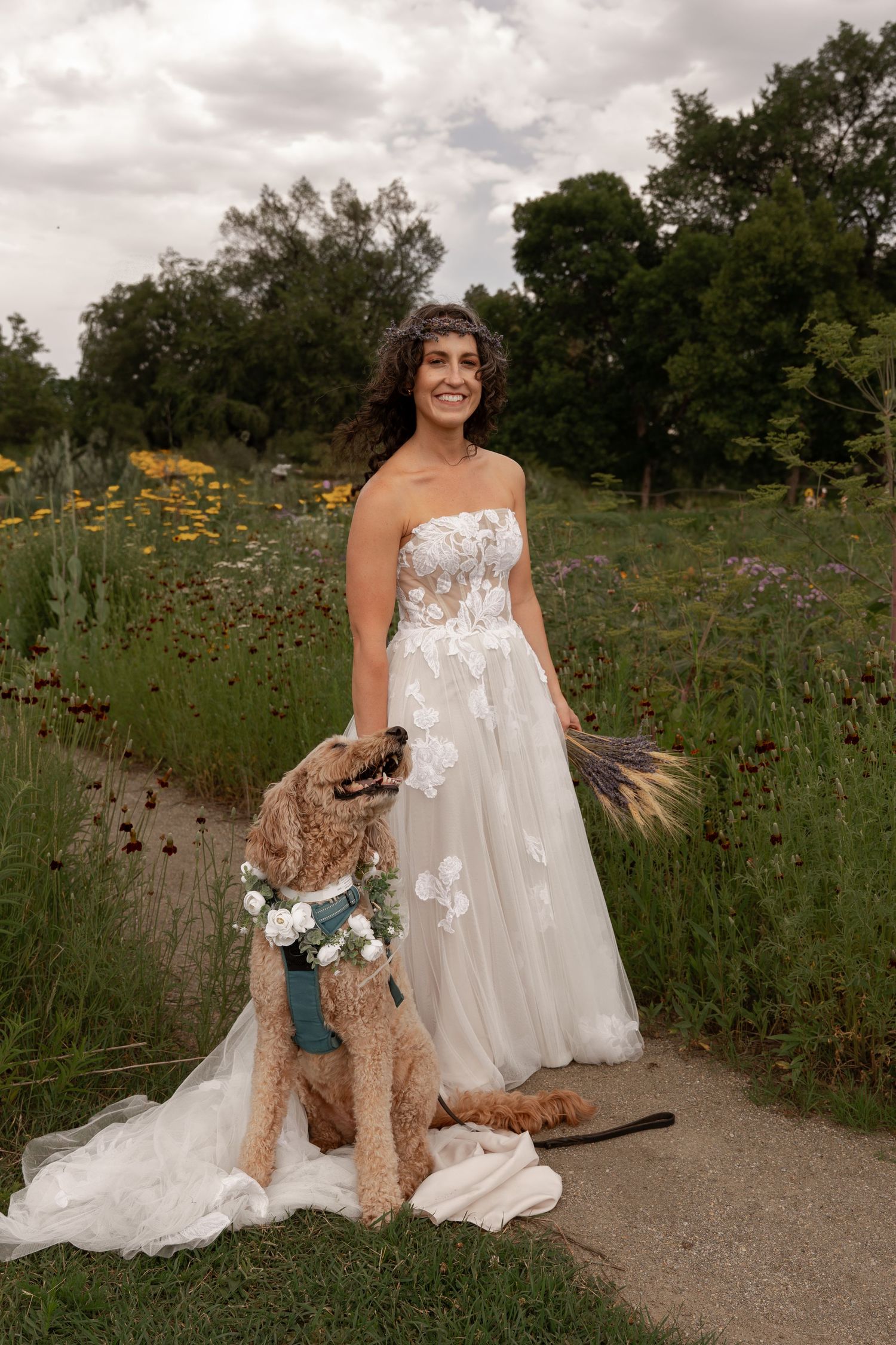 A person in a white wedding dress poses with a brown dog in a field of wildflowers on a path.