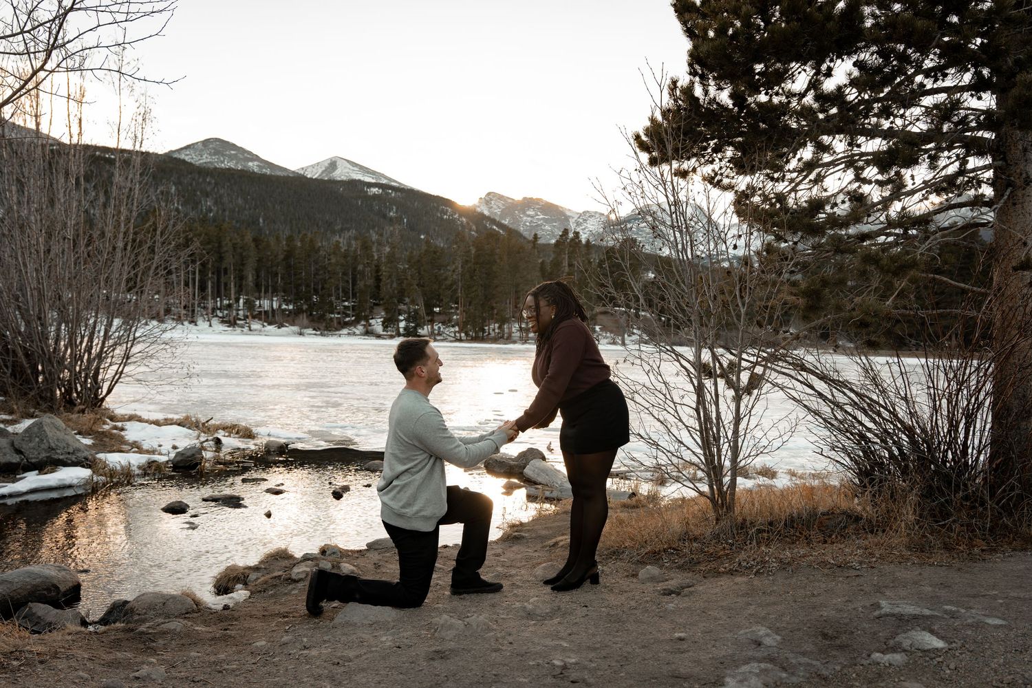 A proposal scene with one person kneeling by a mountain lake in winter.