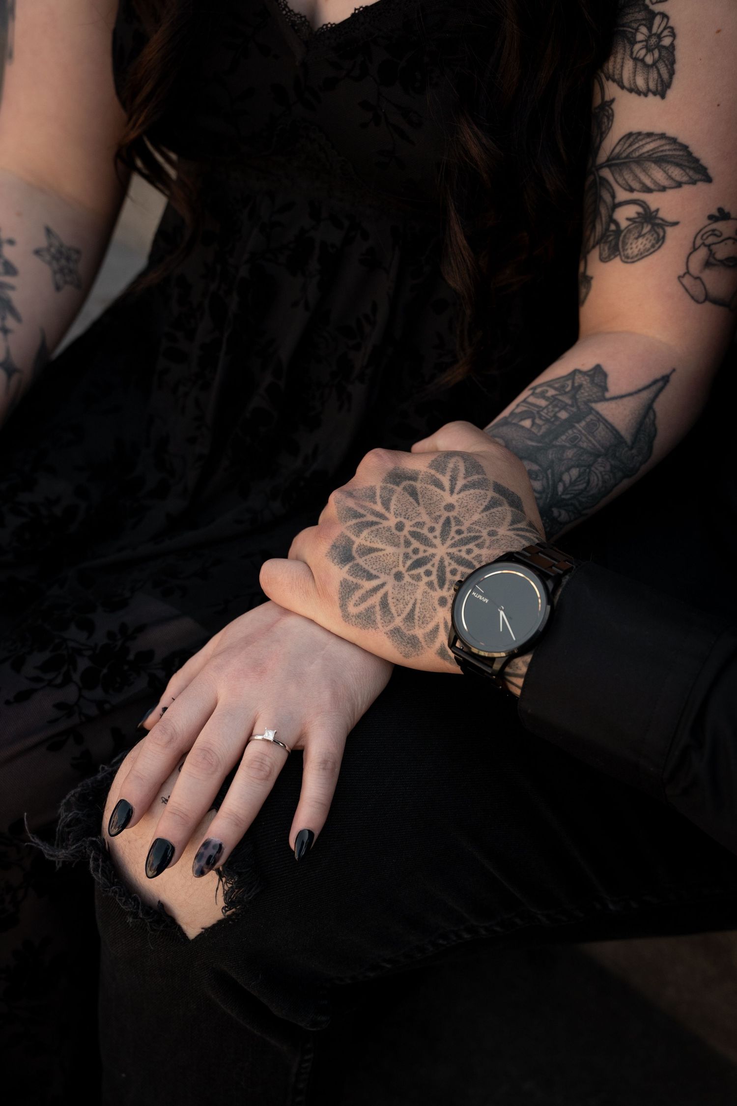 Tattooed woman's arms crossed on lap showcasing floral tattoos, black nails, engagement ring, and black watch.