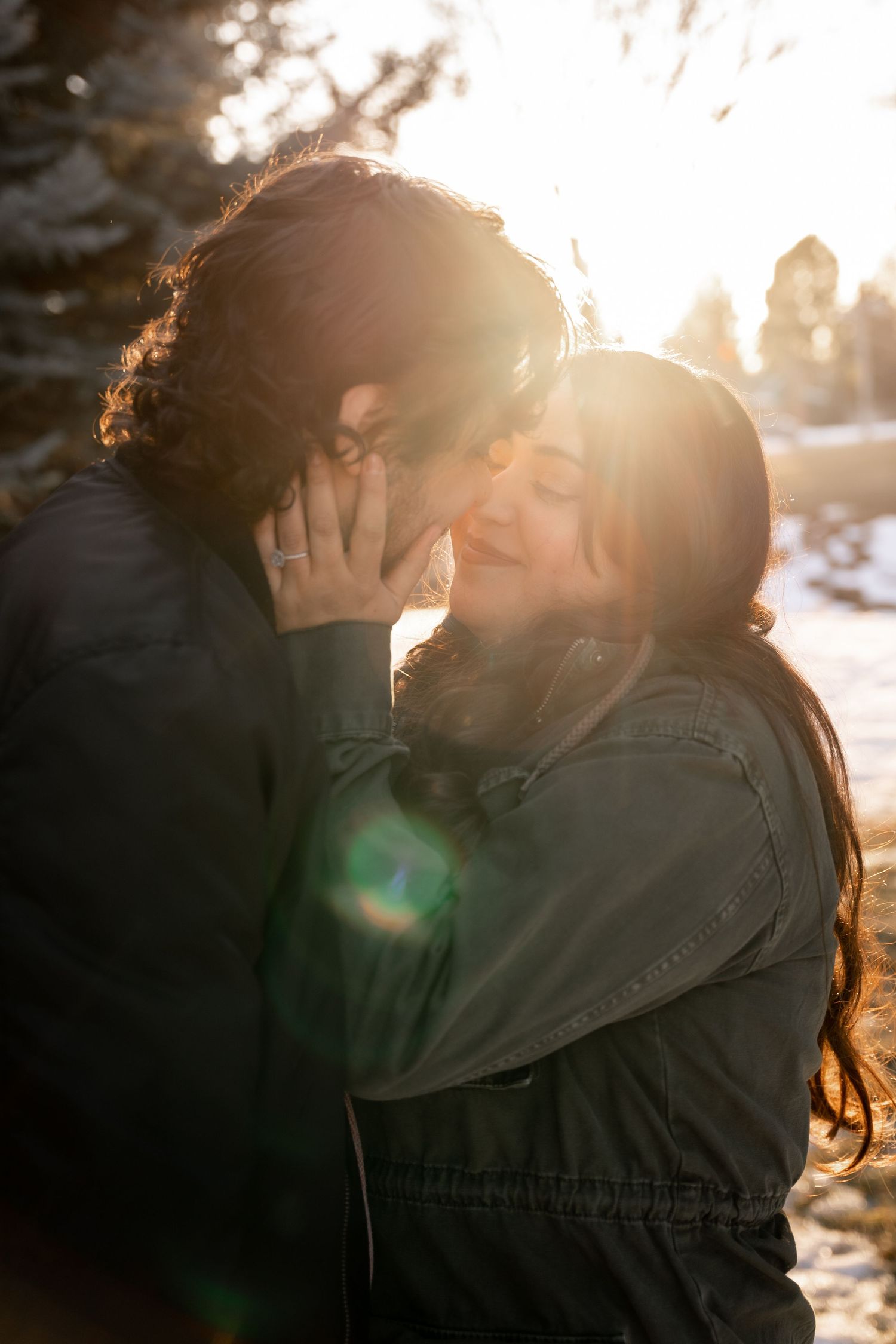 Couple sharing a tender kiss backlit by stunning golden sun flare in a snowy outdoor setting.