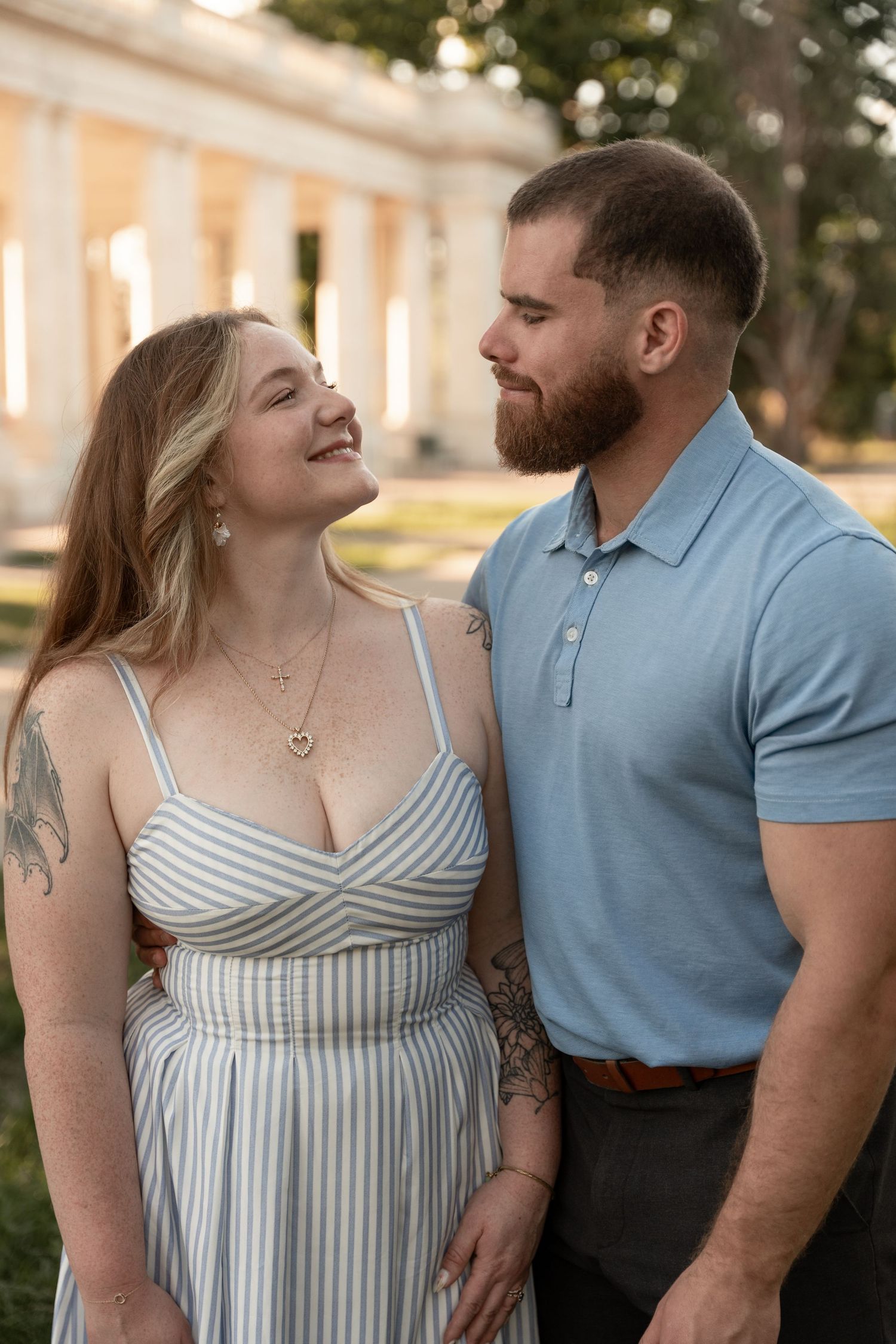 A smiling couple in casual attire share a romantic moment outdoors near white columns at golden hour.