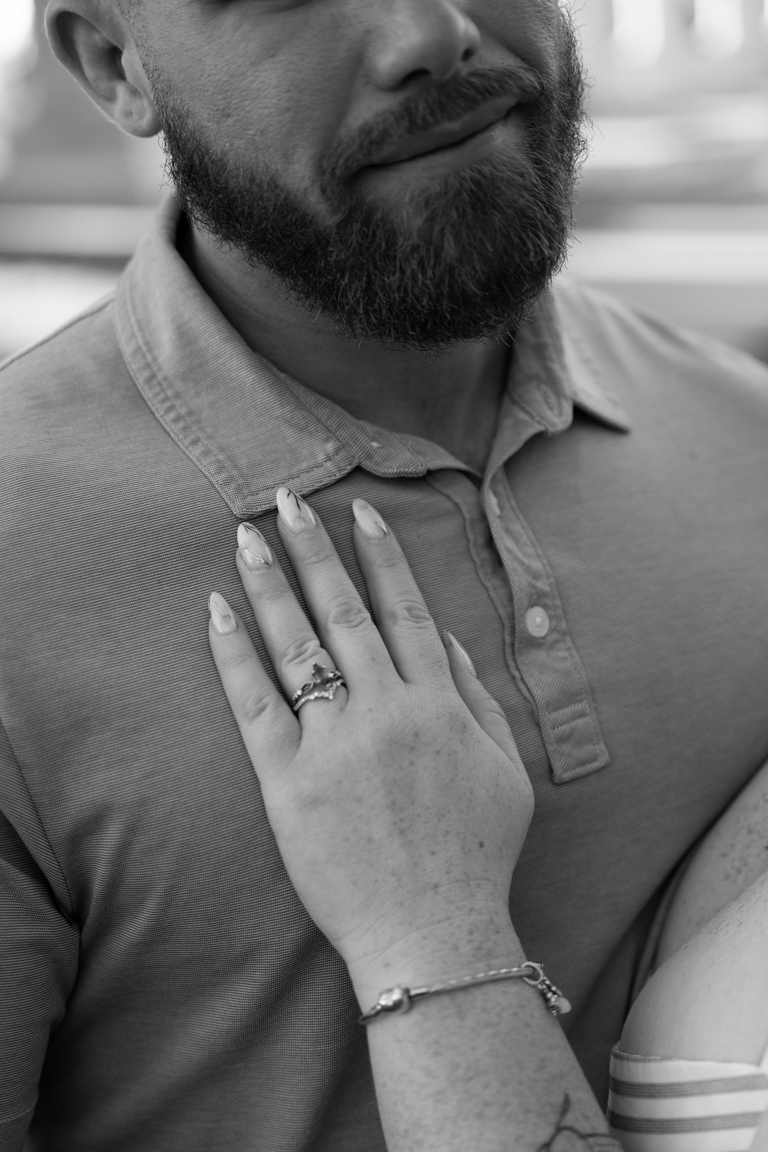 Black and white photo of a woman's hand with rings and bracelet resting on a bearded man's chest.