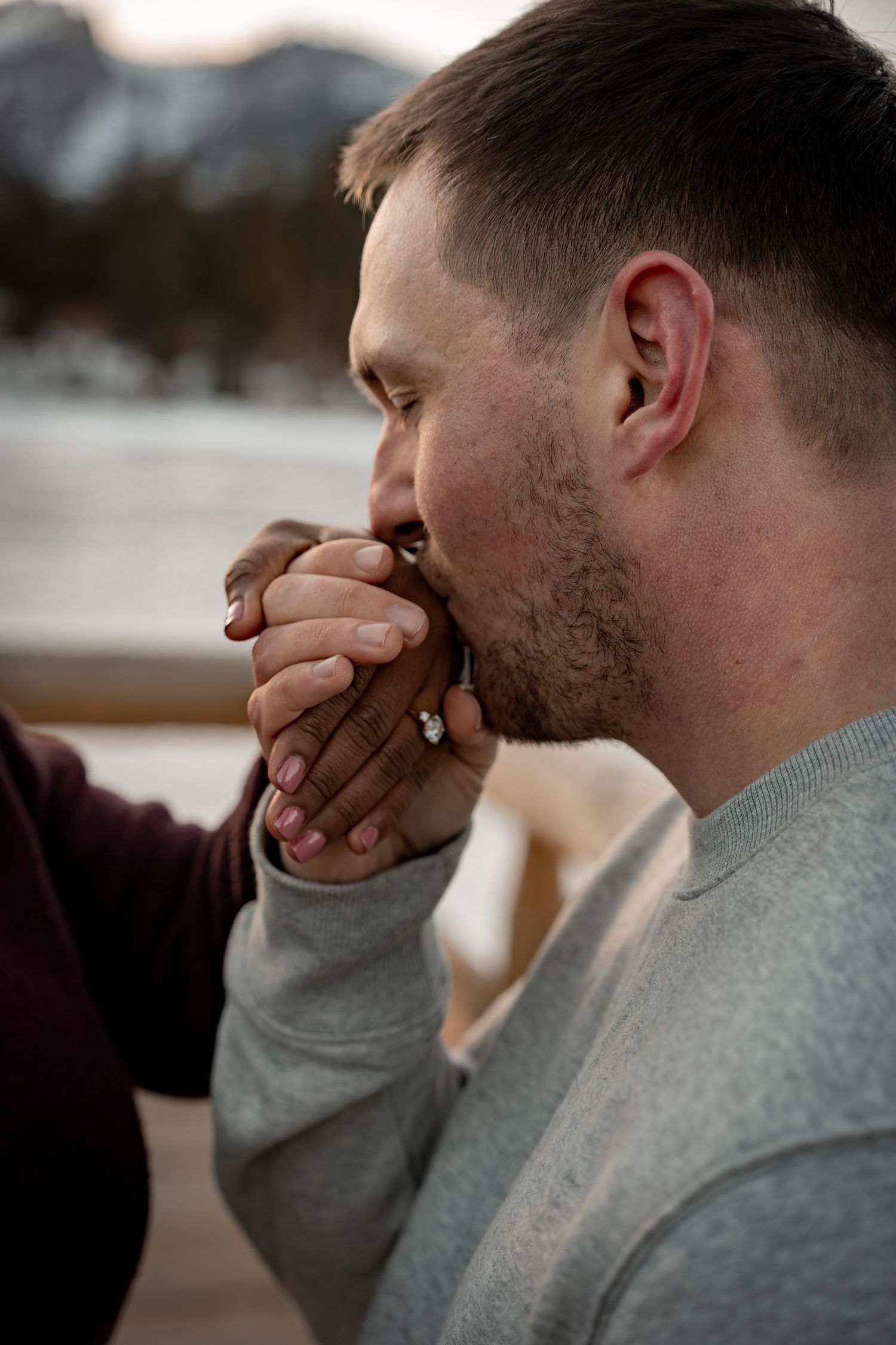 Man gently kissing woman's hand with engagement ring in romantic winter mountain setting.