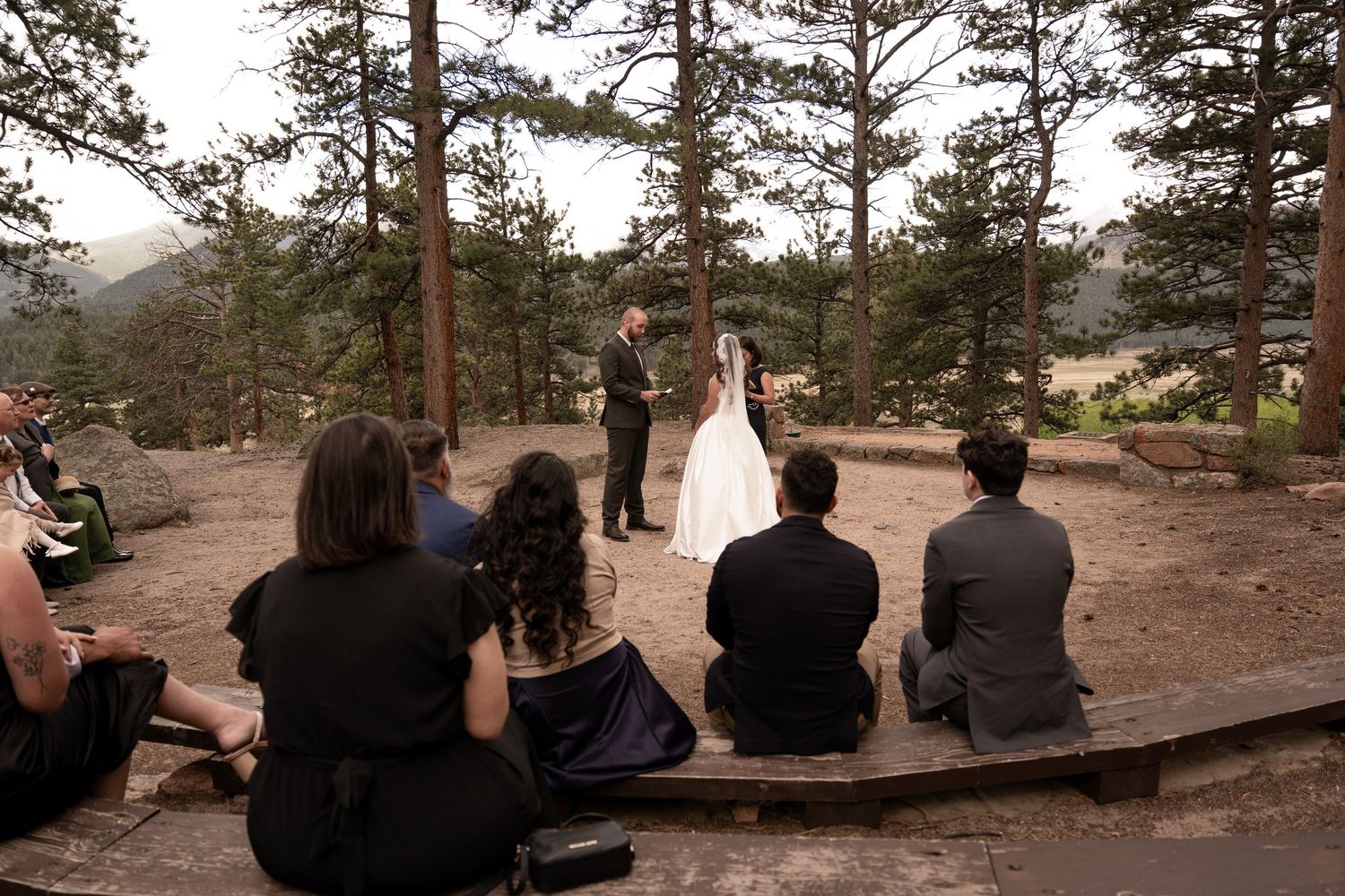 Outdoor wedding ceremony in a pine forest with guests seated on rocks watching couple exchange vows.