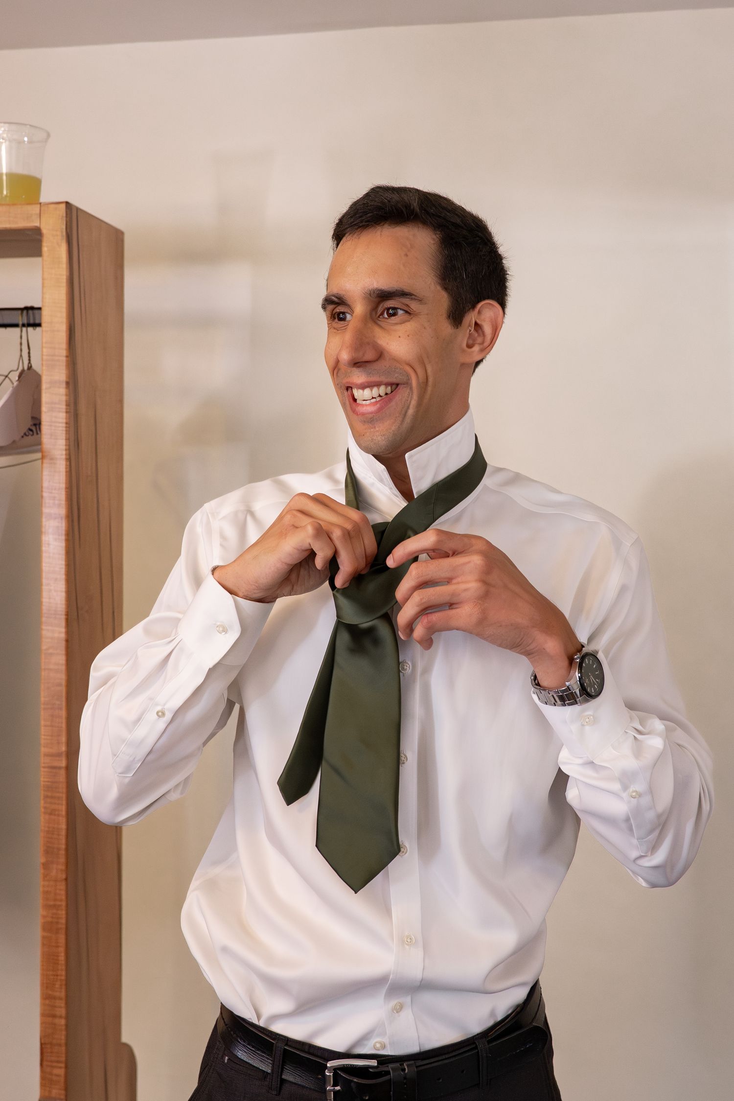 Groom smiles while putting on his dark green tie getting ready for his wedding.