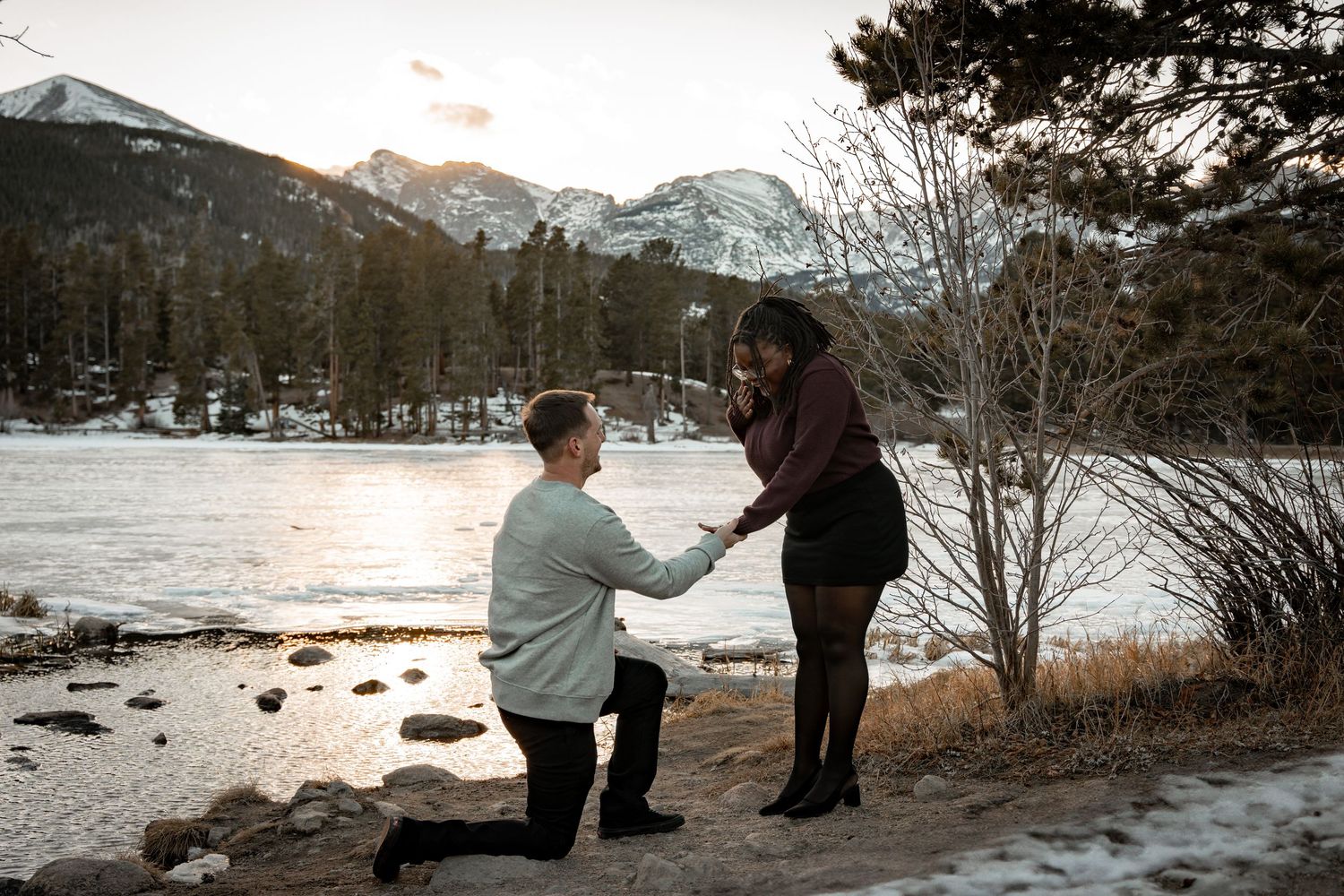 Man proposes to woman on one knee by a frozen mountain lake at sunset in a snowy winter landscape.