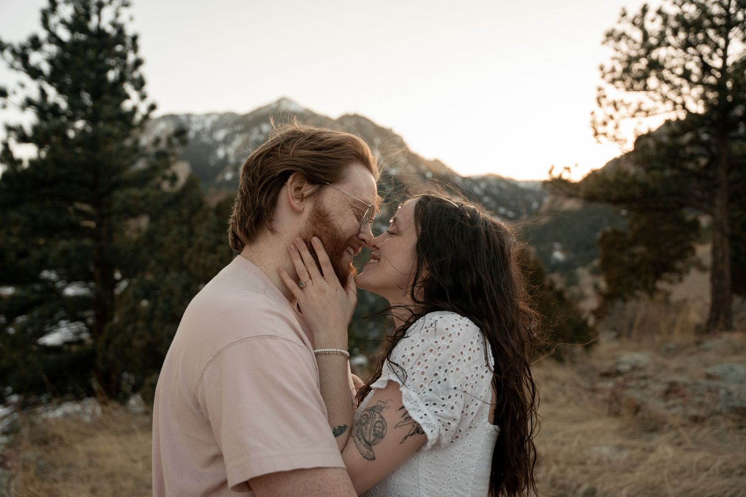 Couple sharing a romantic kiss at sunset with mountain and pine tree backdrop in a scenic outdoor setting.