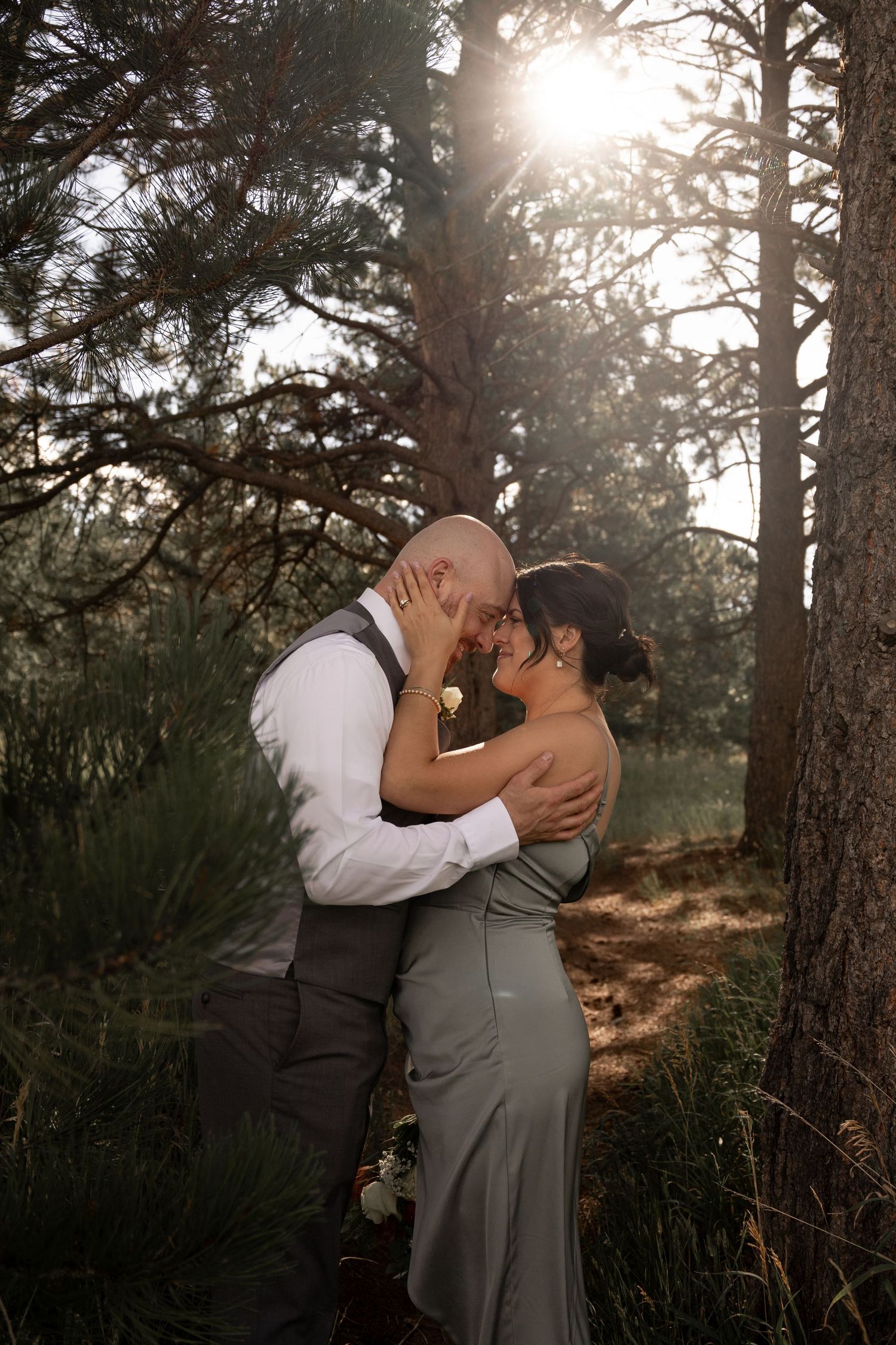 A romantic wedding kiss captured in a sunlit forest with tall pine trees creating a magical backdrop.