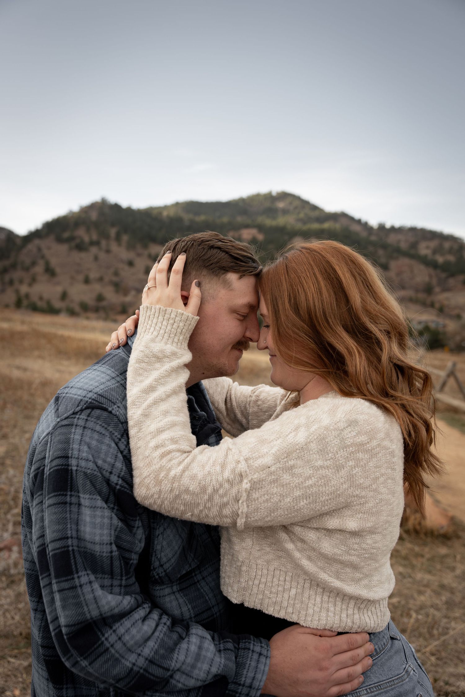 Couple embracing forehead-to-forehead in a mountain meadow, woman in cream sweater with red hair, man in plaid shirt.