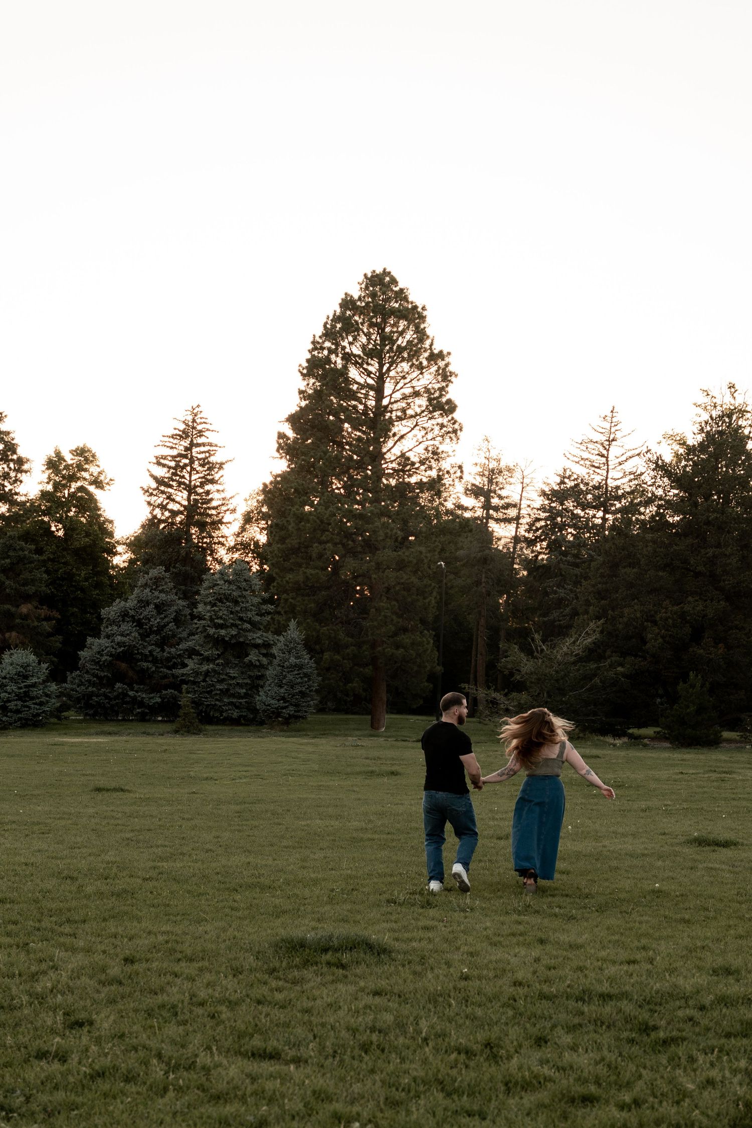 Couple holding hands running through a green park field at sunset with tall evergreen trees in the background.