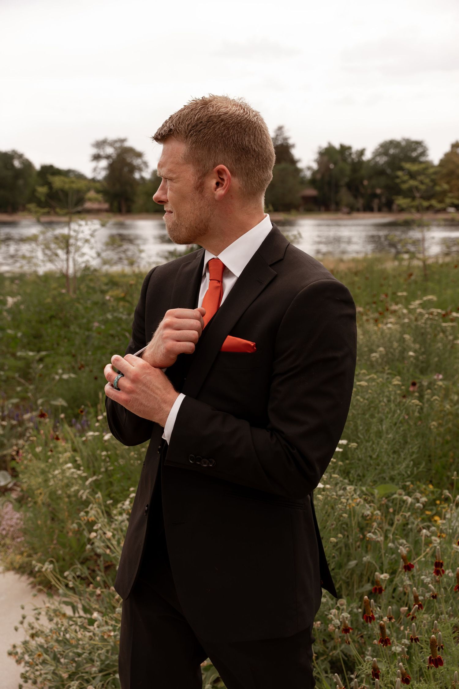 Man in black suit with red tie adjusts his collar while standing in a wildflower field near a lake.