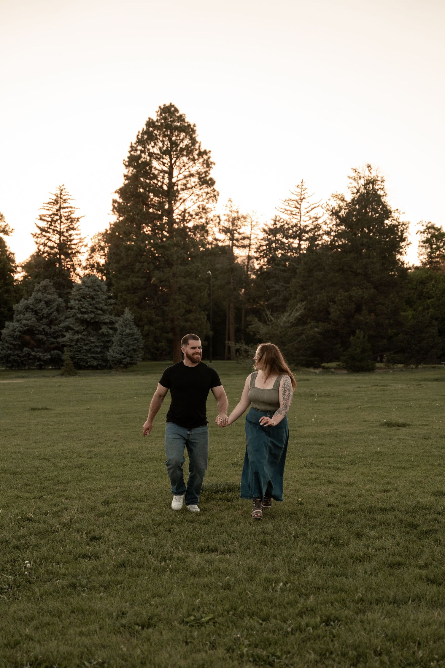 Couple holding hands walking through a green field at golden hour with tall trees in the background.