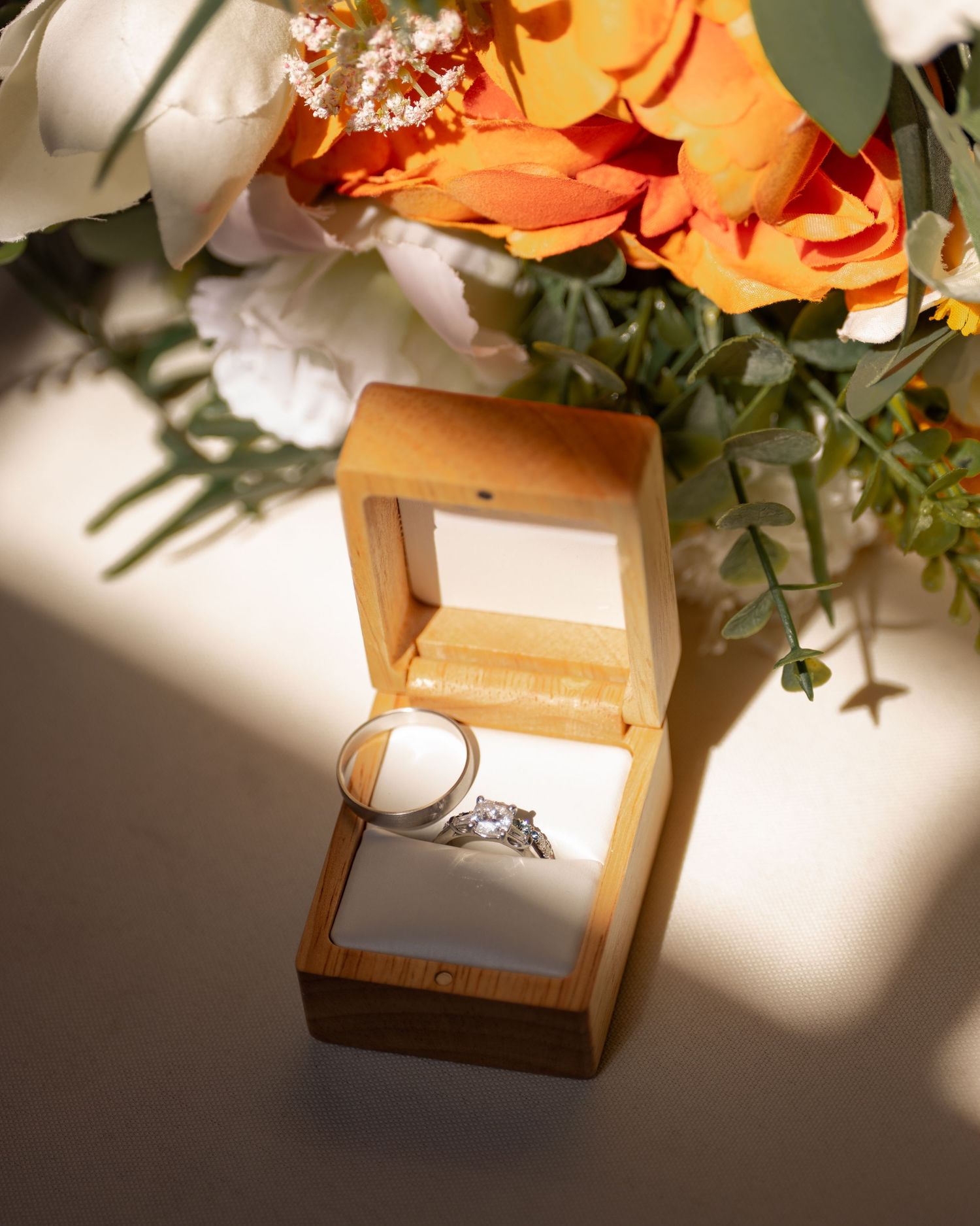 Wedding ring in wooden box with orange and white flowers casting shadows.