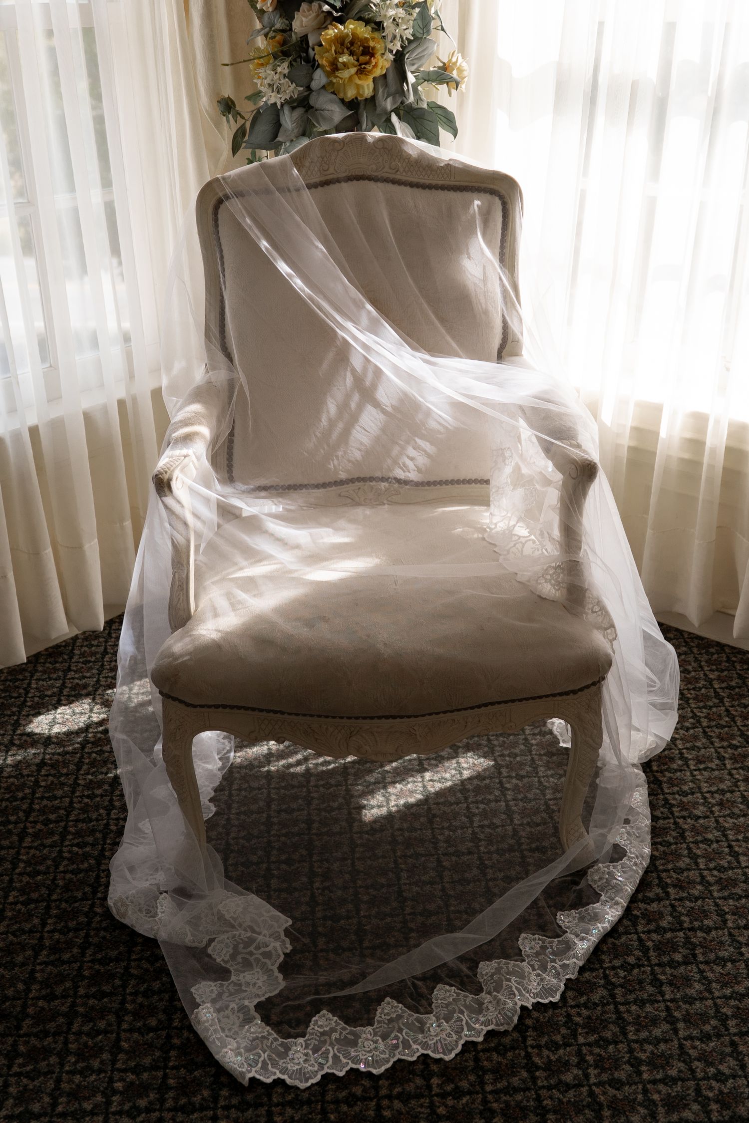 Wedding dress with delicate lace veil draped over chair near sunlit window.