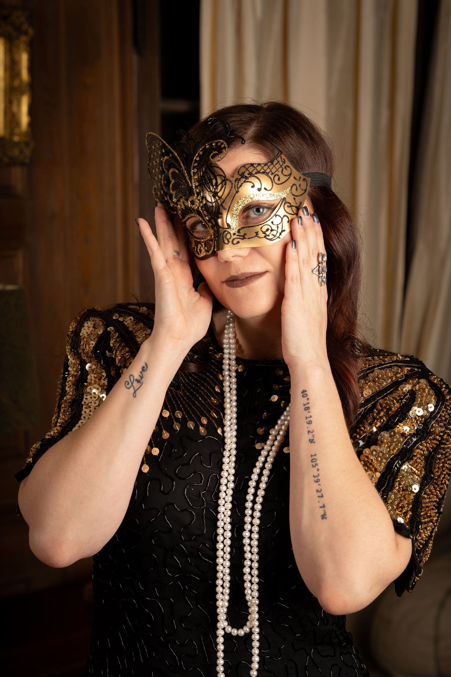 Artistic portrait with masquerade mask and pearl necklace against cream curtain backdrop.
