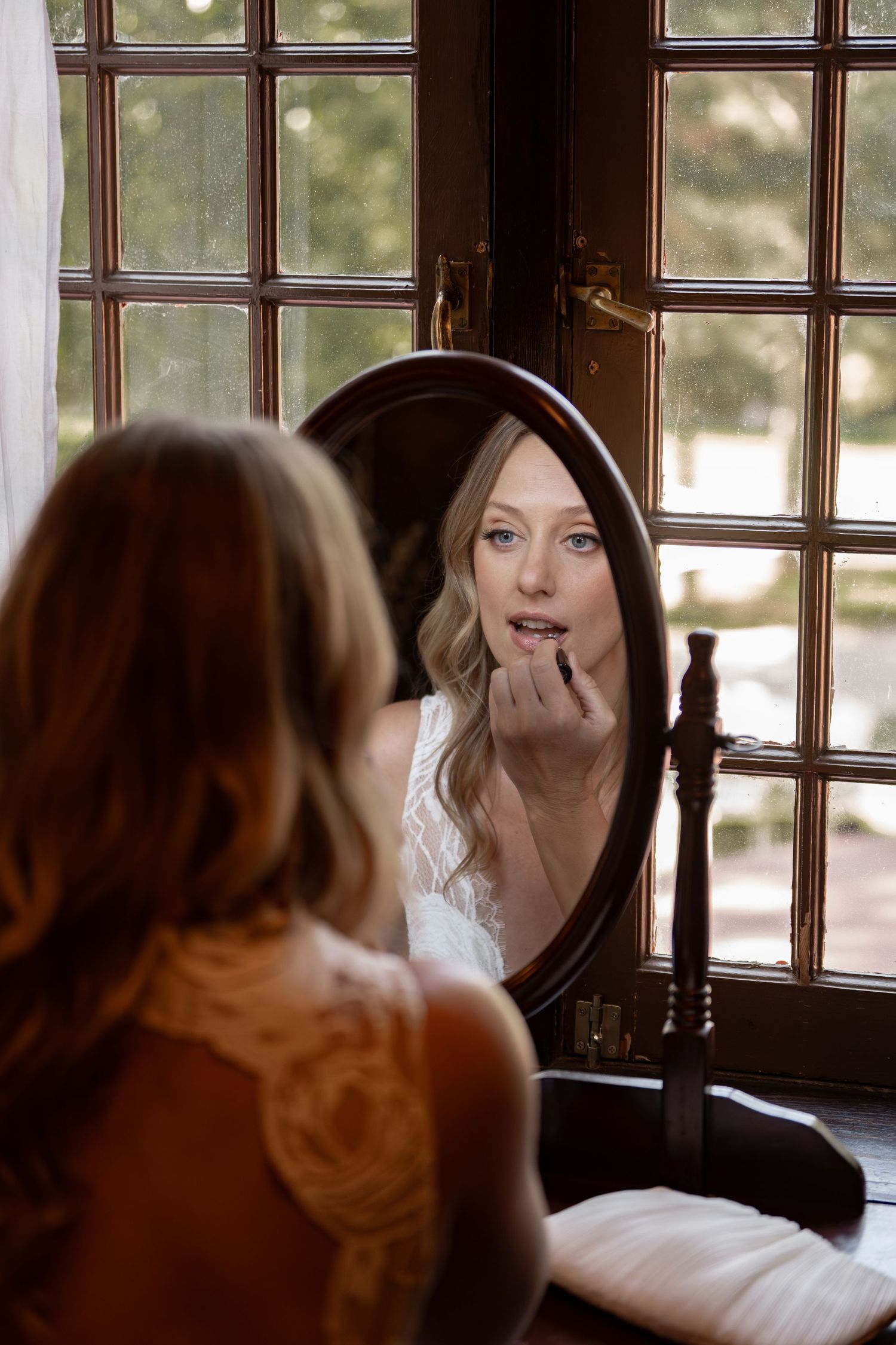 A reflection in a vintage mirror shows a person getting ready near a window with natural light streaming in.