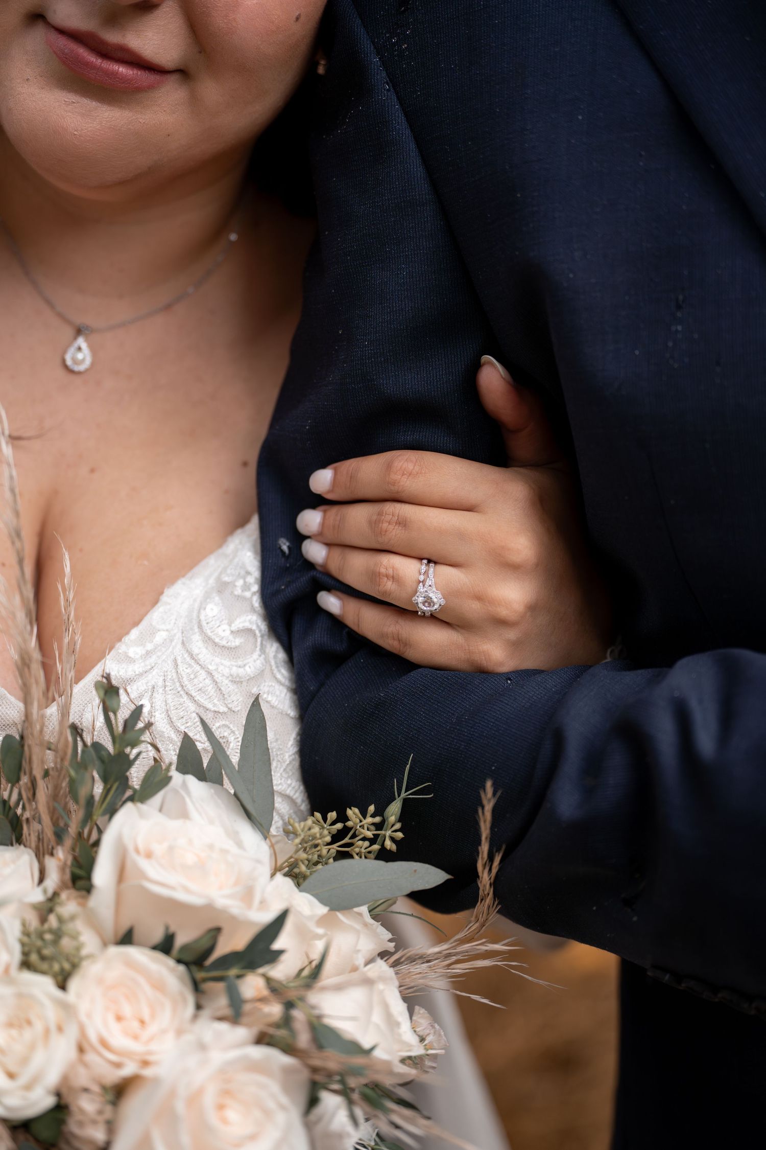 Close-up of wedding bouquet with blush roses and engagement ring detail against navy suit and white lace dress.