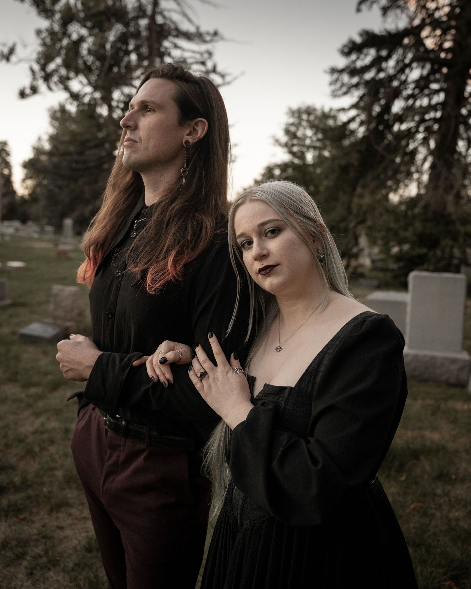 Gothic couple in black attire posing in cemetery at dusk, woman with blonde hair and dark lipstick.