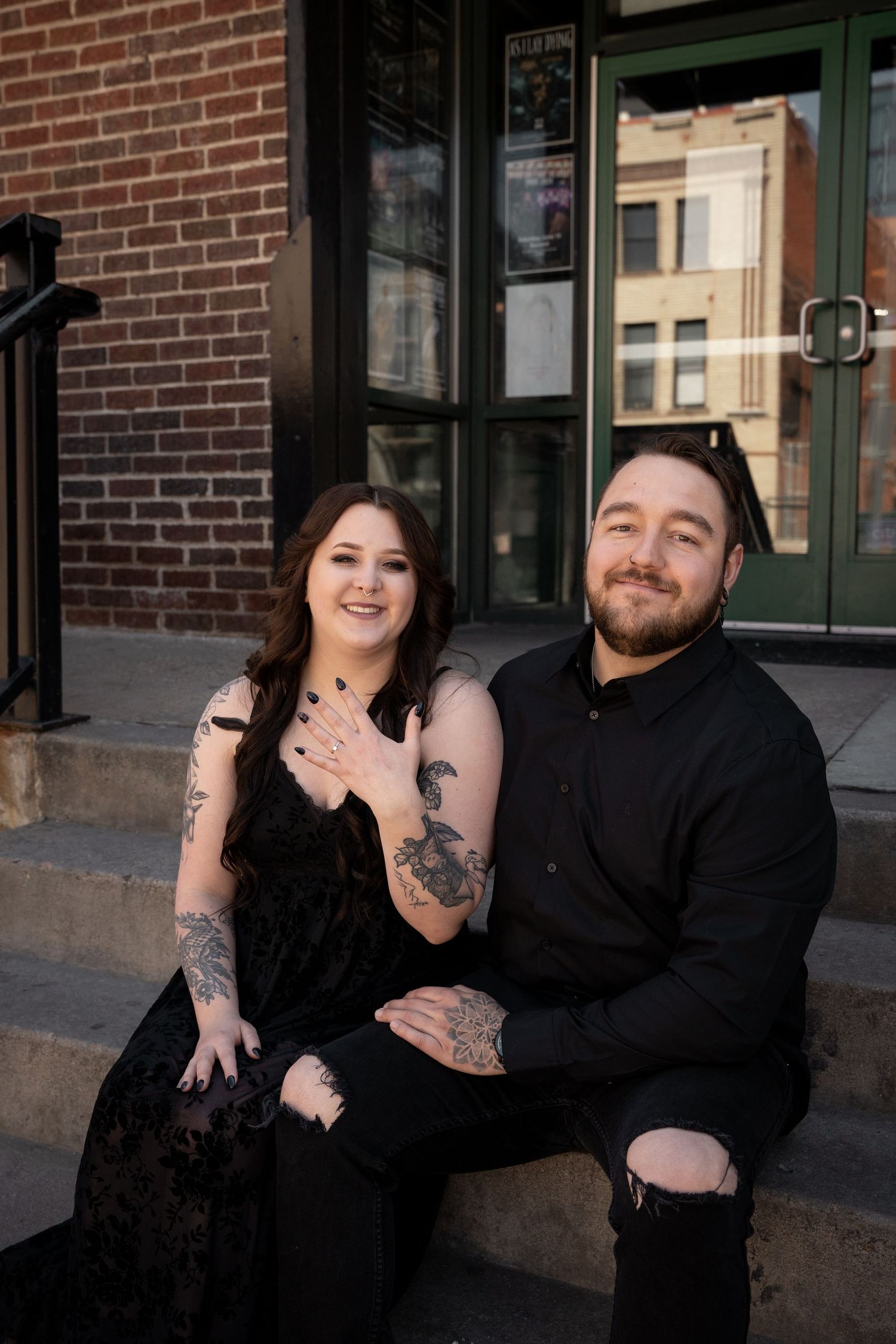 Couple sitting on steps showing engagement ring, both wearing black outfits with tattoos, smiling in front of brick building.