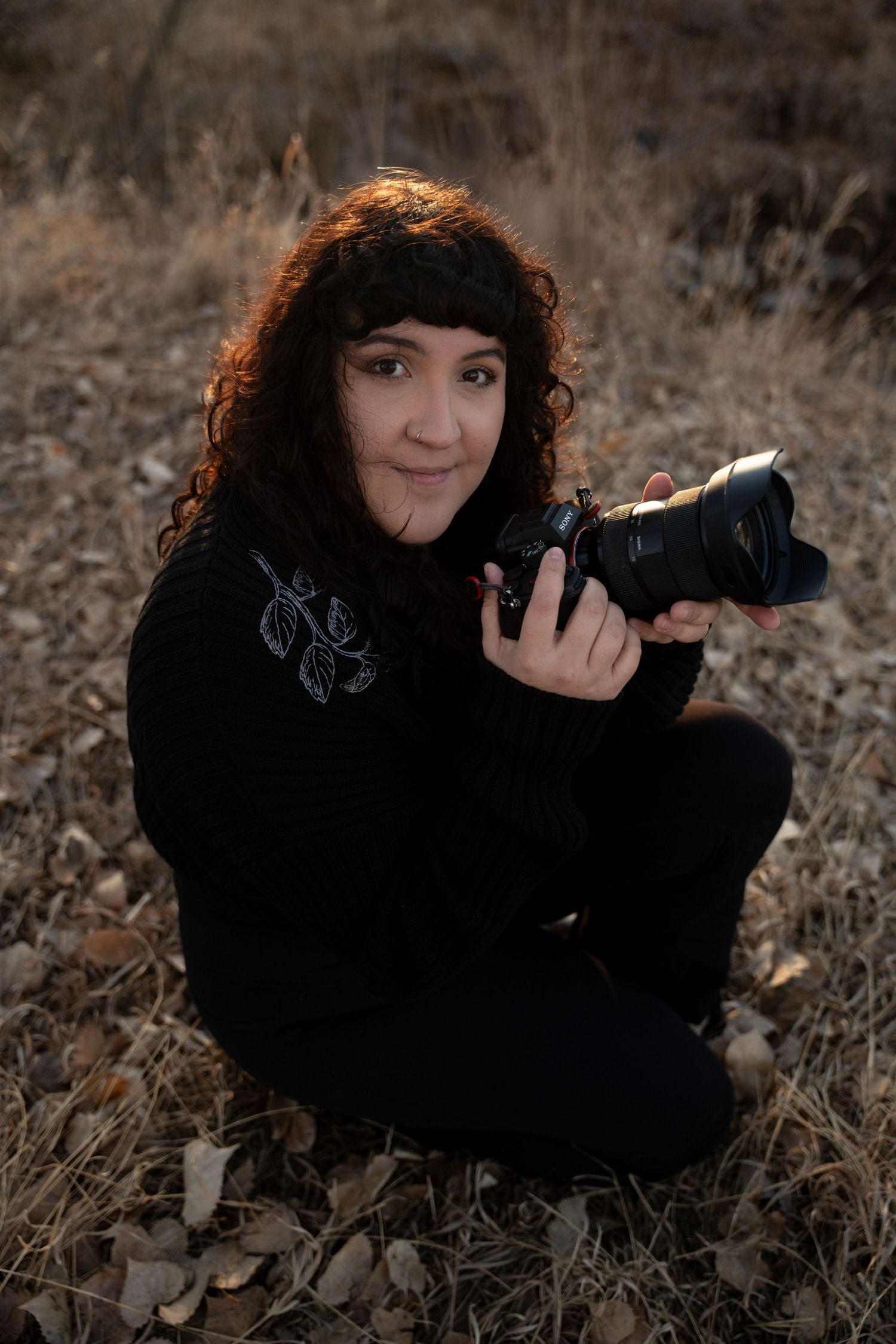 A photographer in black clothing sits with camera equipment in an autumn field.