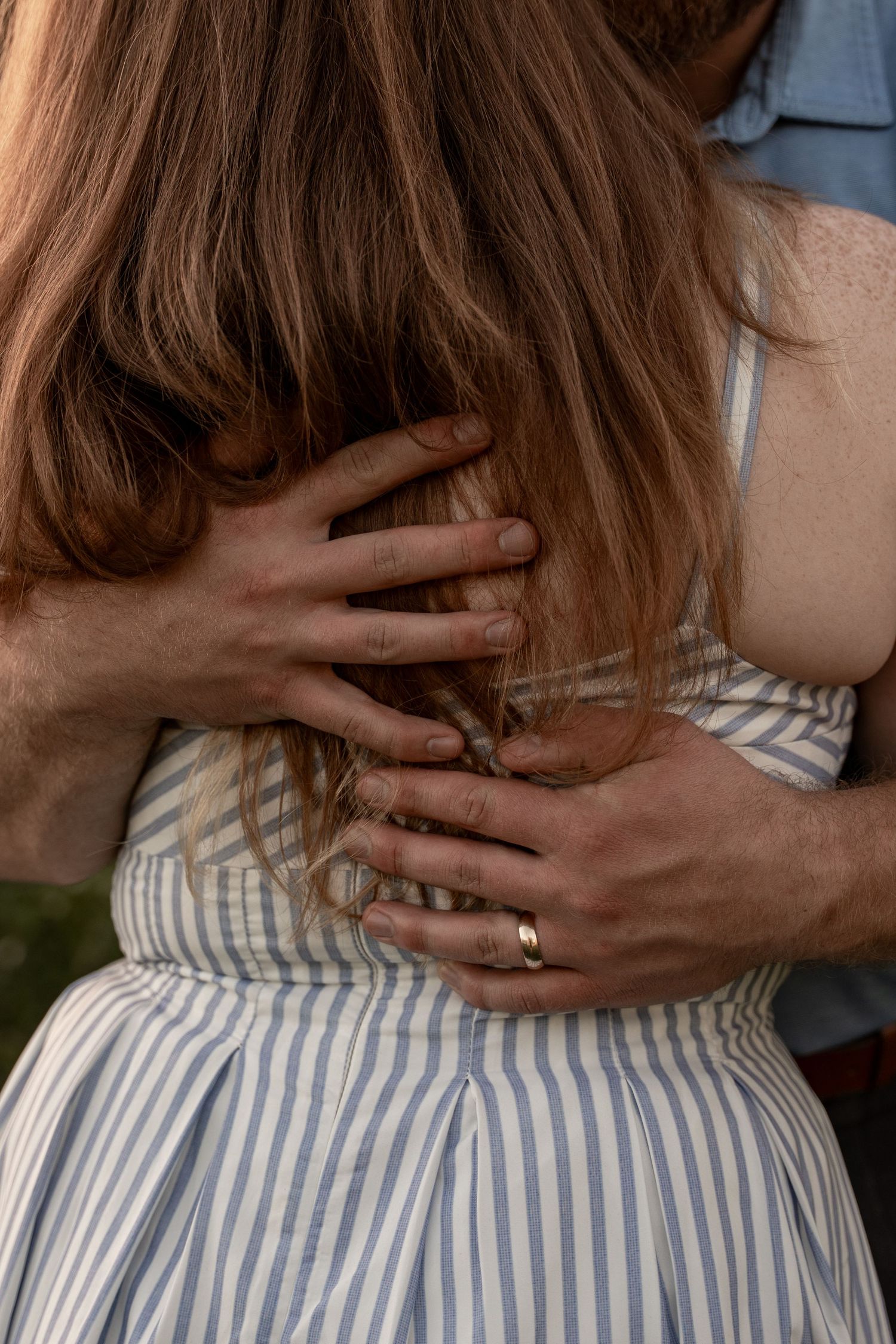 A man with a gold ring embraces a woman in a striped blue dress from behind, her long brown hair cascading down.