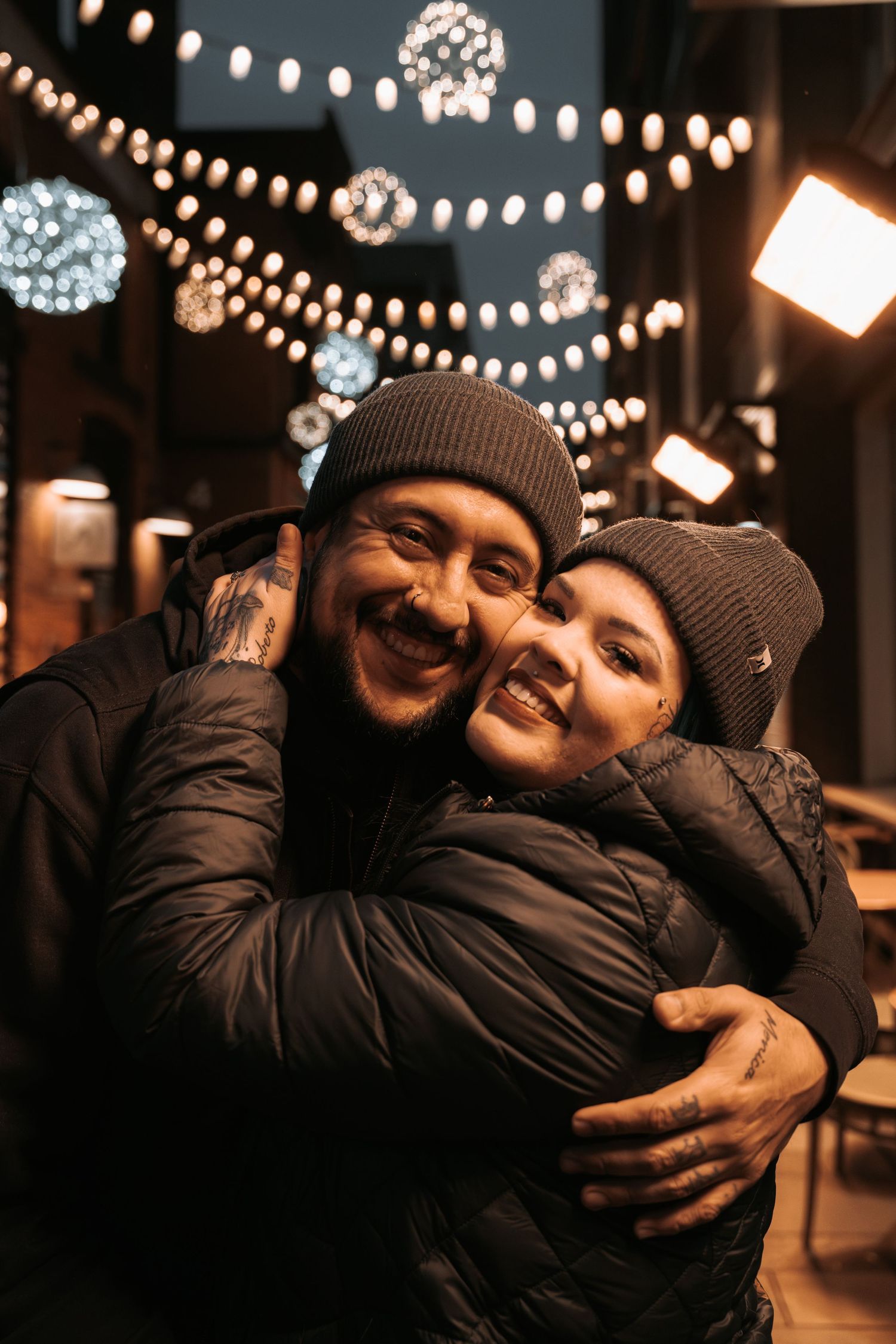 Happy couple hugging outdoors at night under festive string lights, both wearing dark winter jackets and beanies.