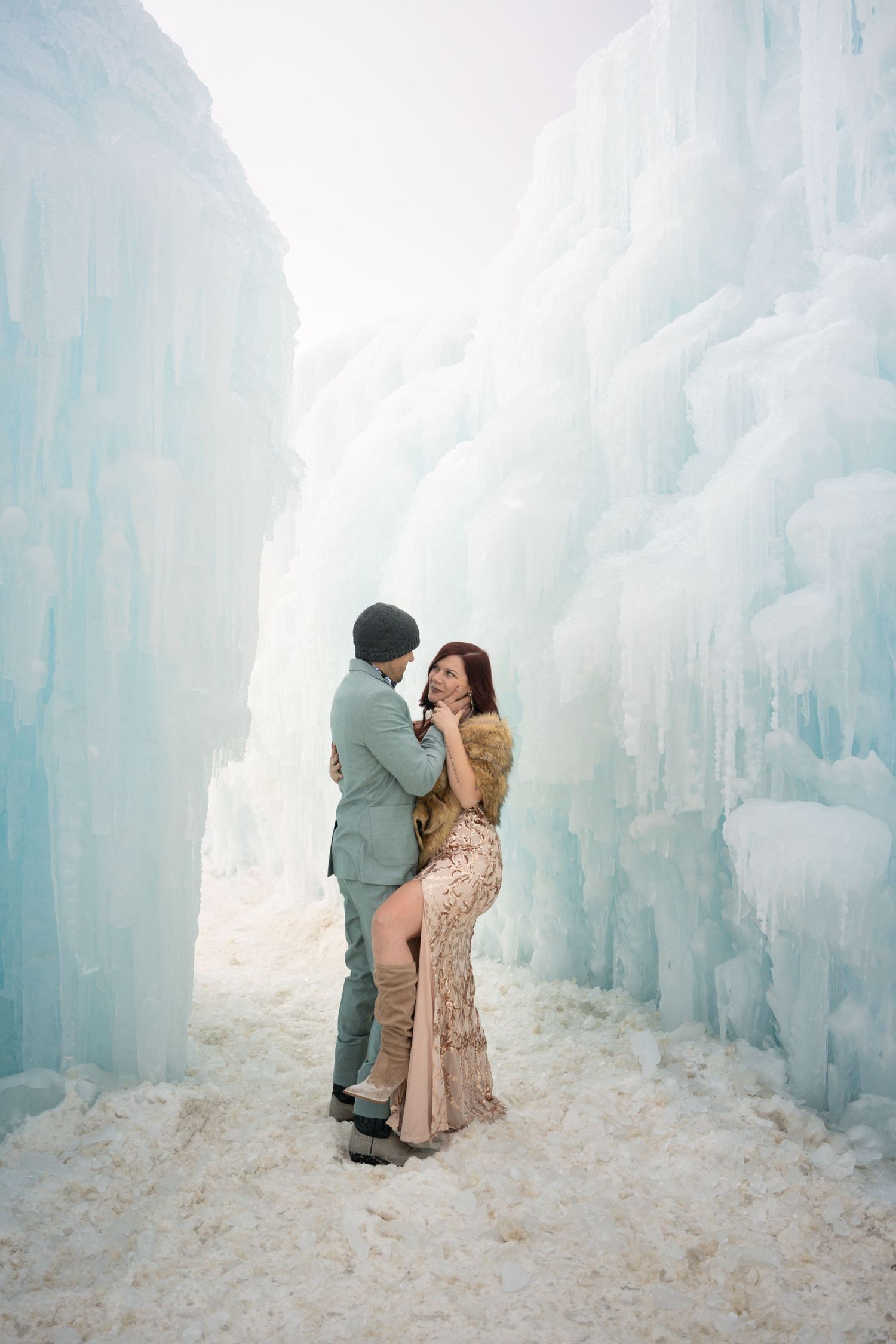 Couple embracing romantically in a stunning ice castle corridor with towering blue ice walls and snow-covered ground.