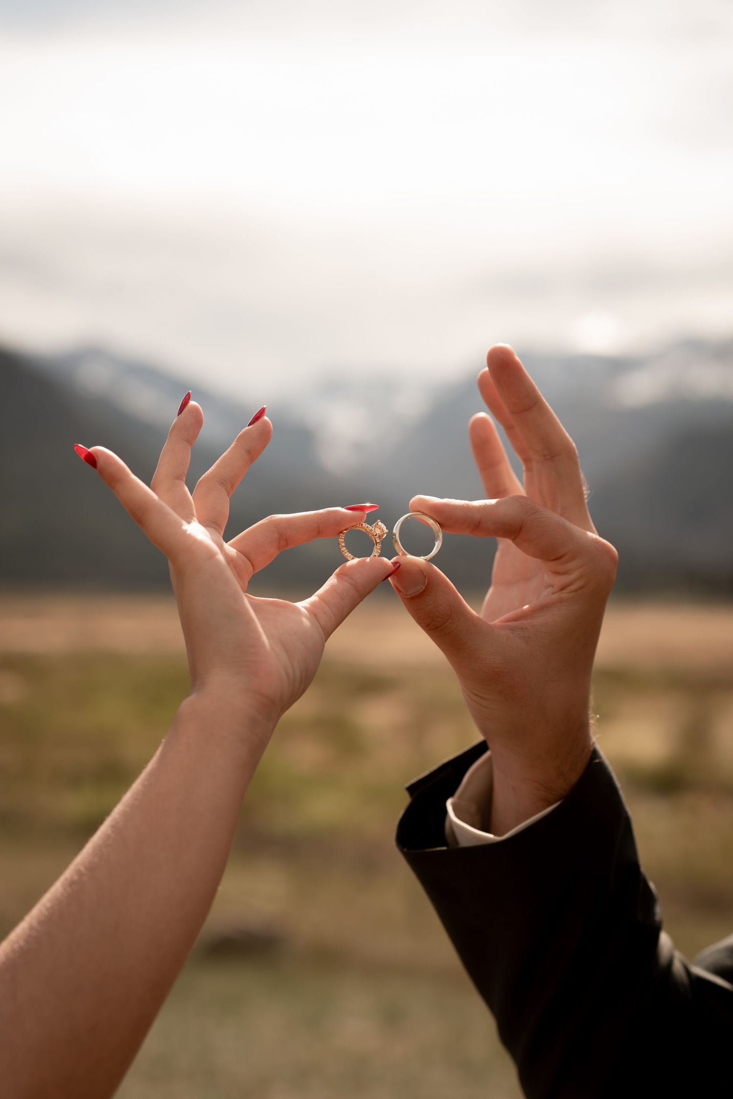 Couple holding two silver wedding rings together against a scenic mountain backdrop, with red-nailed bride and suited groom.