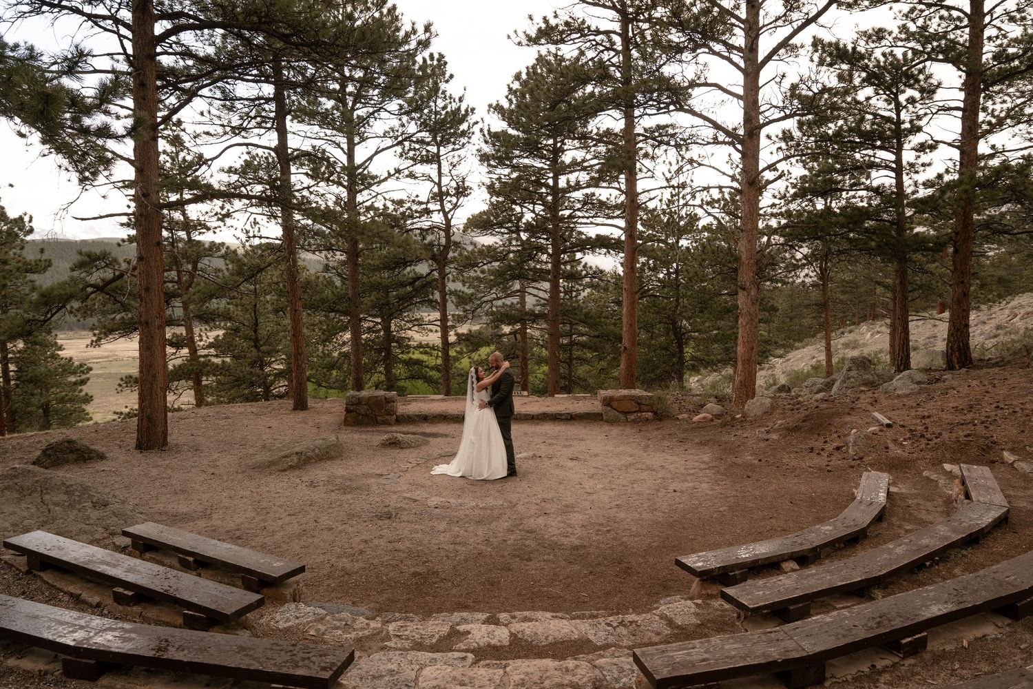 Bride and groom embrace in rustic outdoor amphitheater surrounded by tall pine trees during wedding.