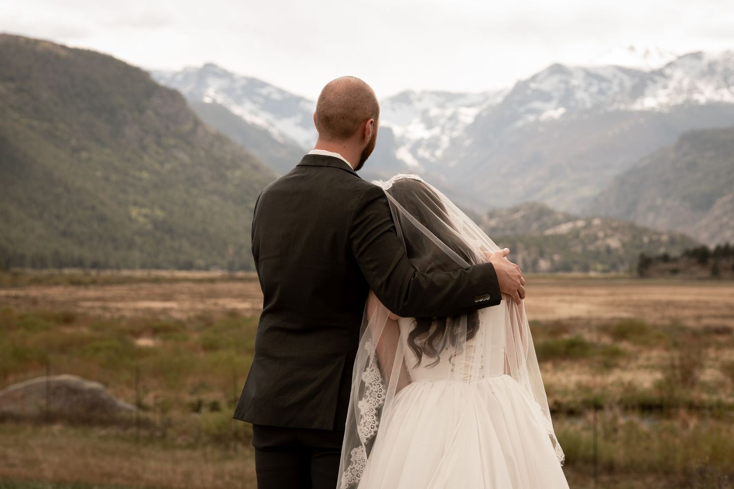 Bride and groom stand together facing snow-capped mountains in a scenic outdoor wedding photo.