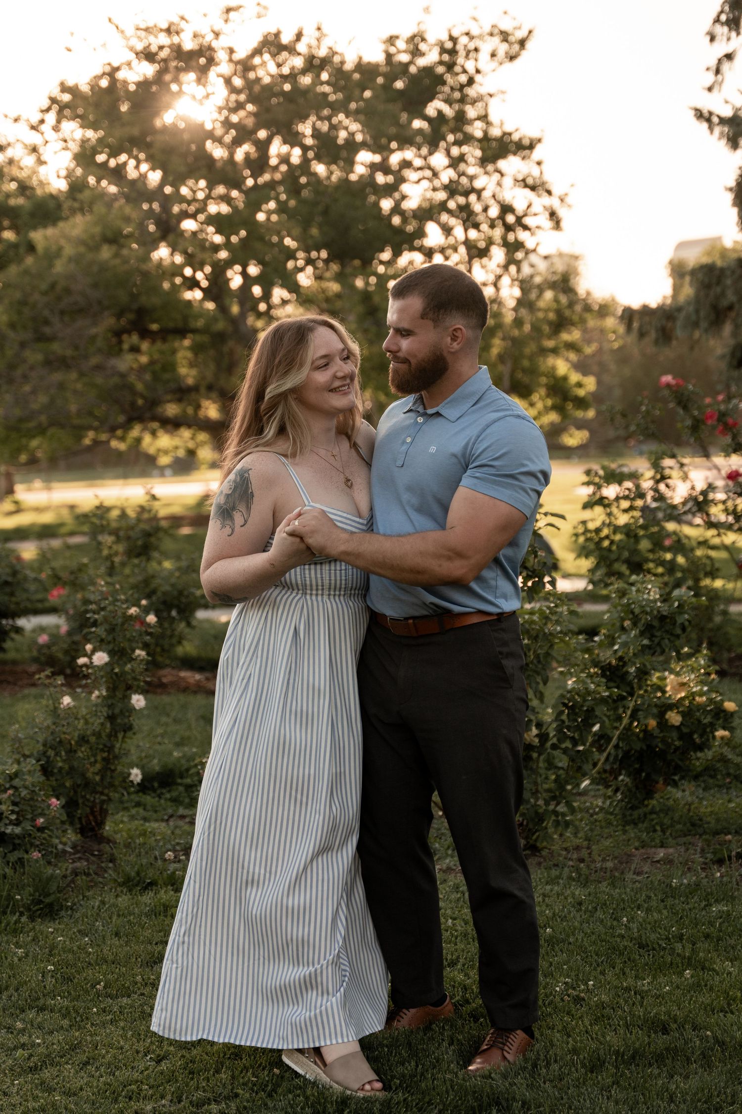 Couple embracing in a lush garden at golden hour, woman in striped dress with tattoo, man in blue polo shirt.