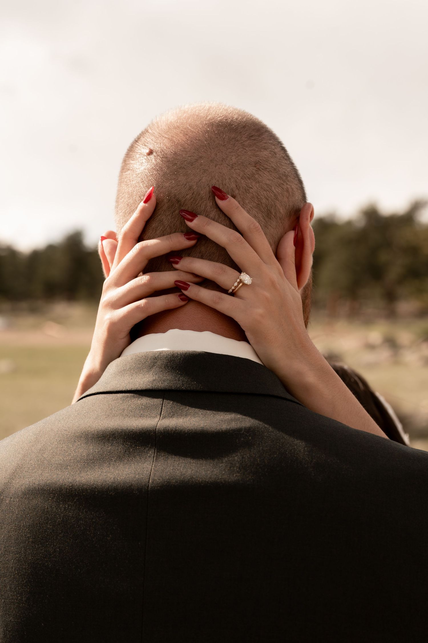 Bride with red nails and diamond engagement ring tenderly holds groom's head during outdoor wedding photo.