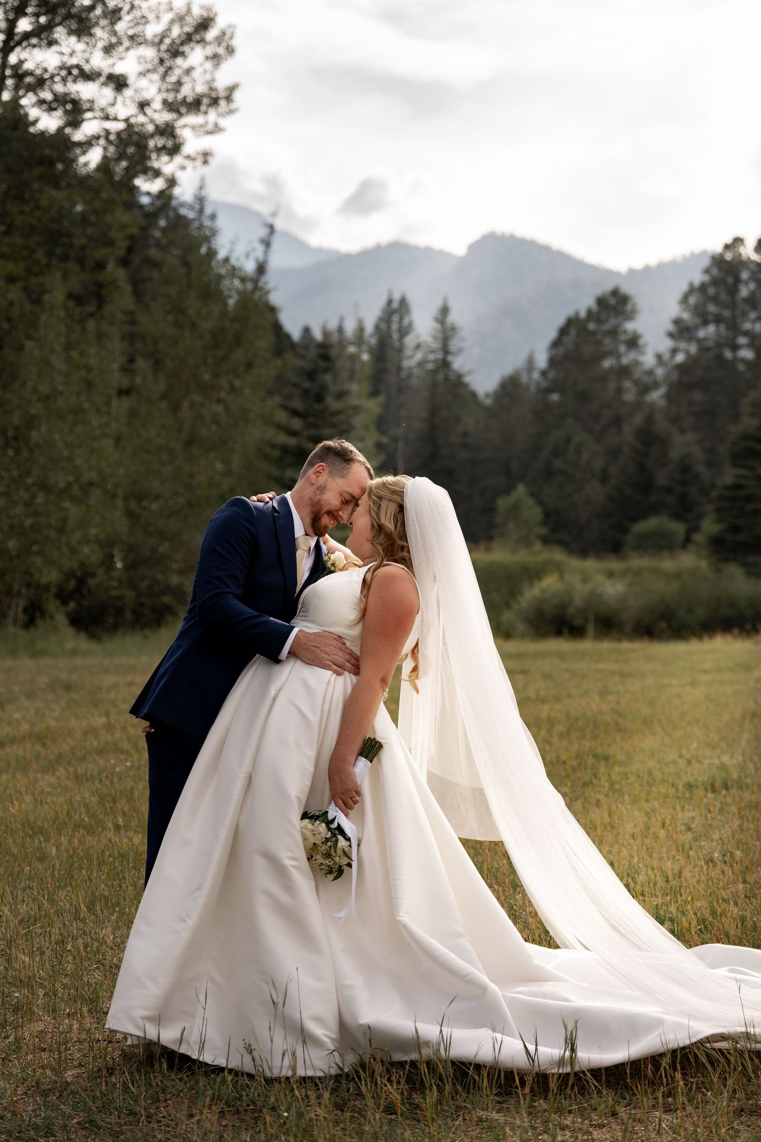 A romantic wedding portrait in a mountain meadow with evergreen trees and misty peaks in the background.