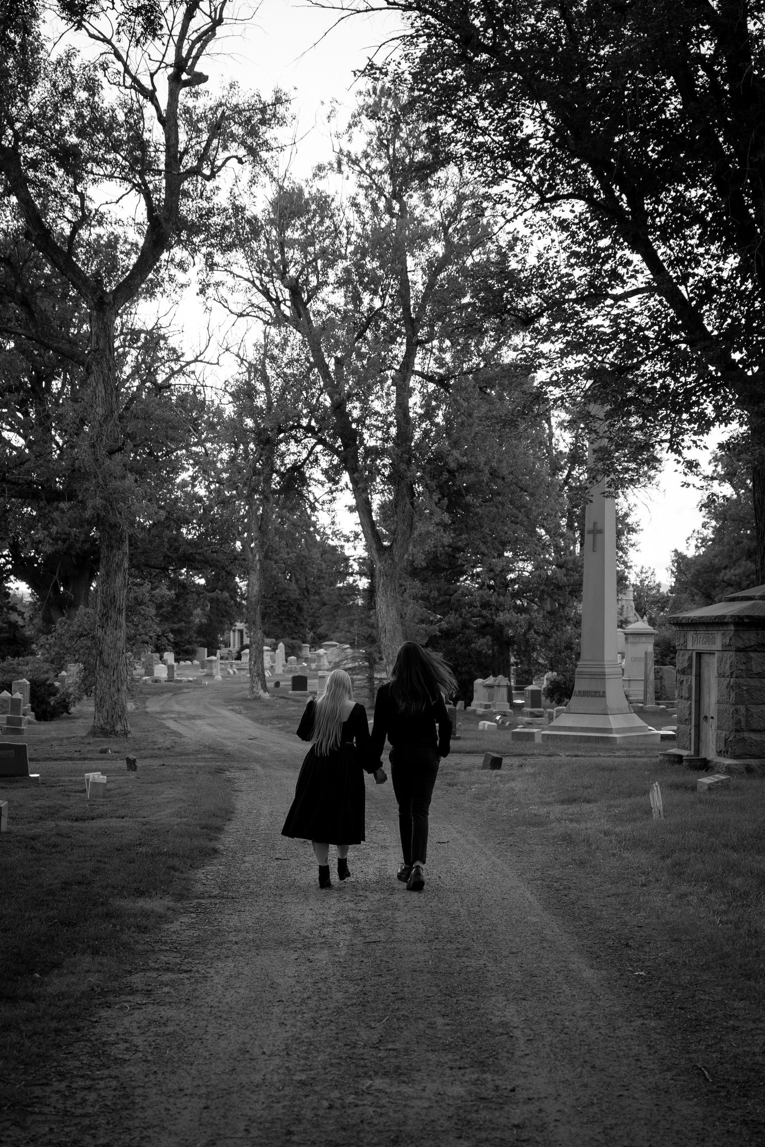 A black and white photograph of two people walking down a cemetery path lined with monuments.