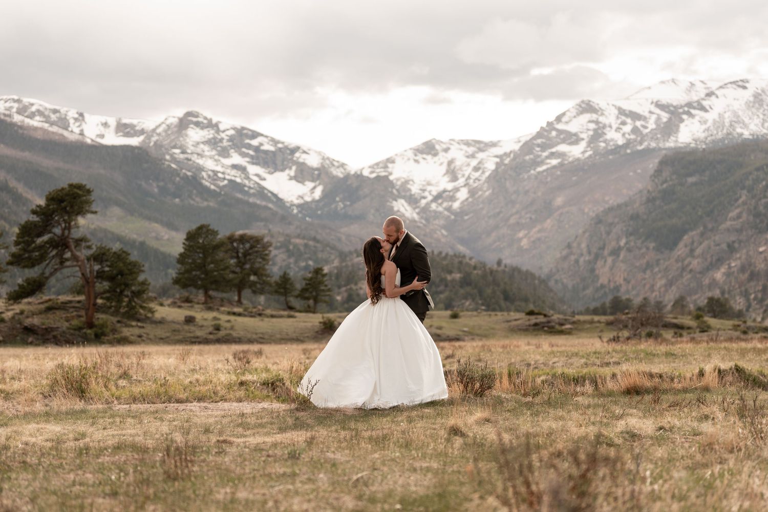Bride and groom share a romantic dip kiss in a mountain meadow with snow-capped Rocky Mountains backdrop.