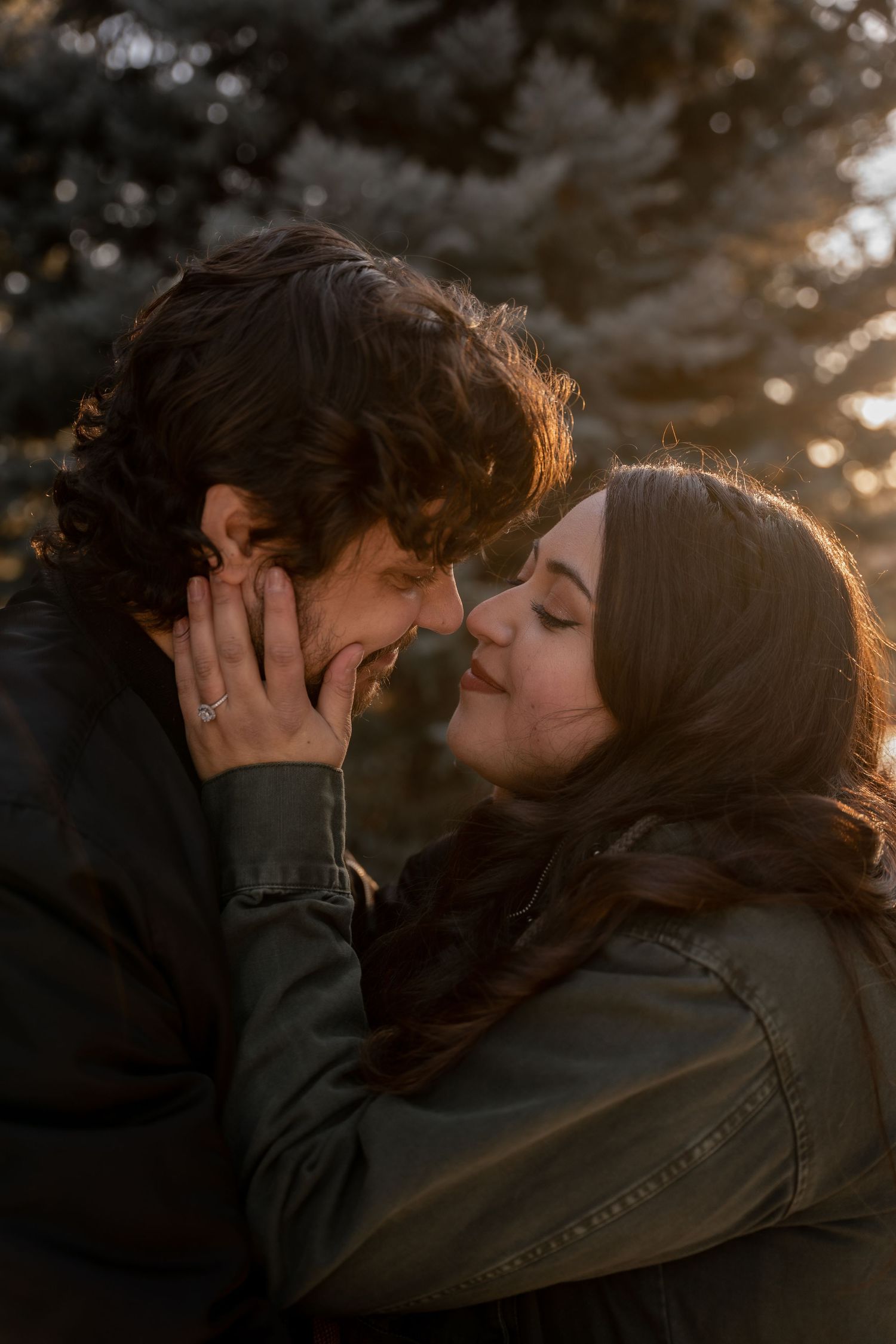 Couple sharing a romantic almost-kiss moment outdoors near evergreen trees in warm golden light.