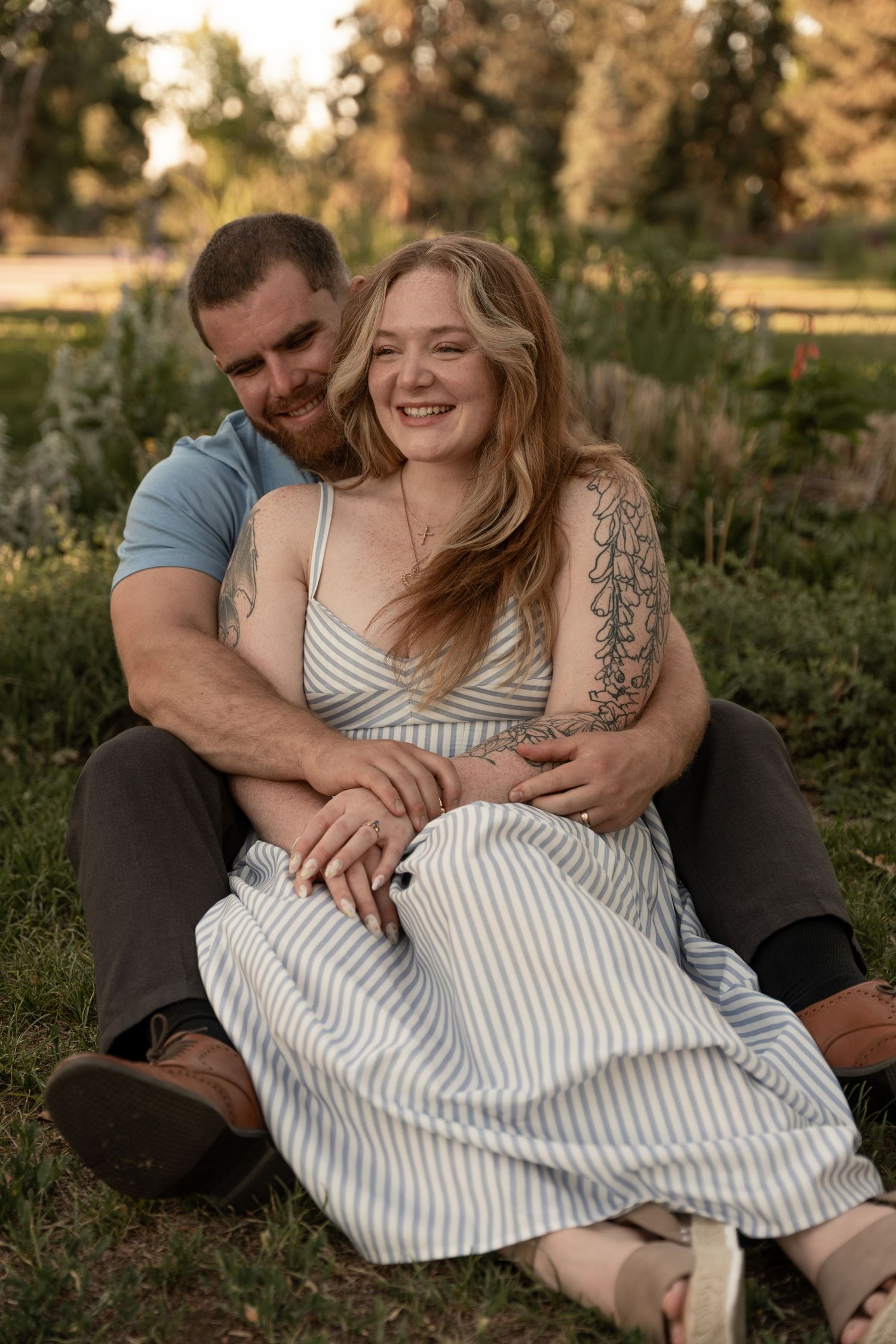 Couple sitting on grass laughing together in a park at sunset, woman in striped dress.
