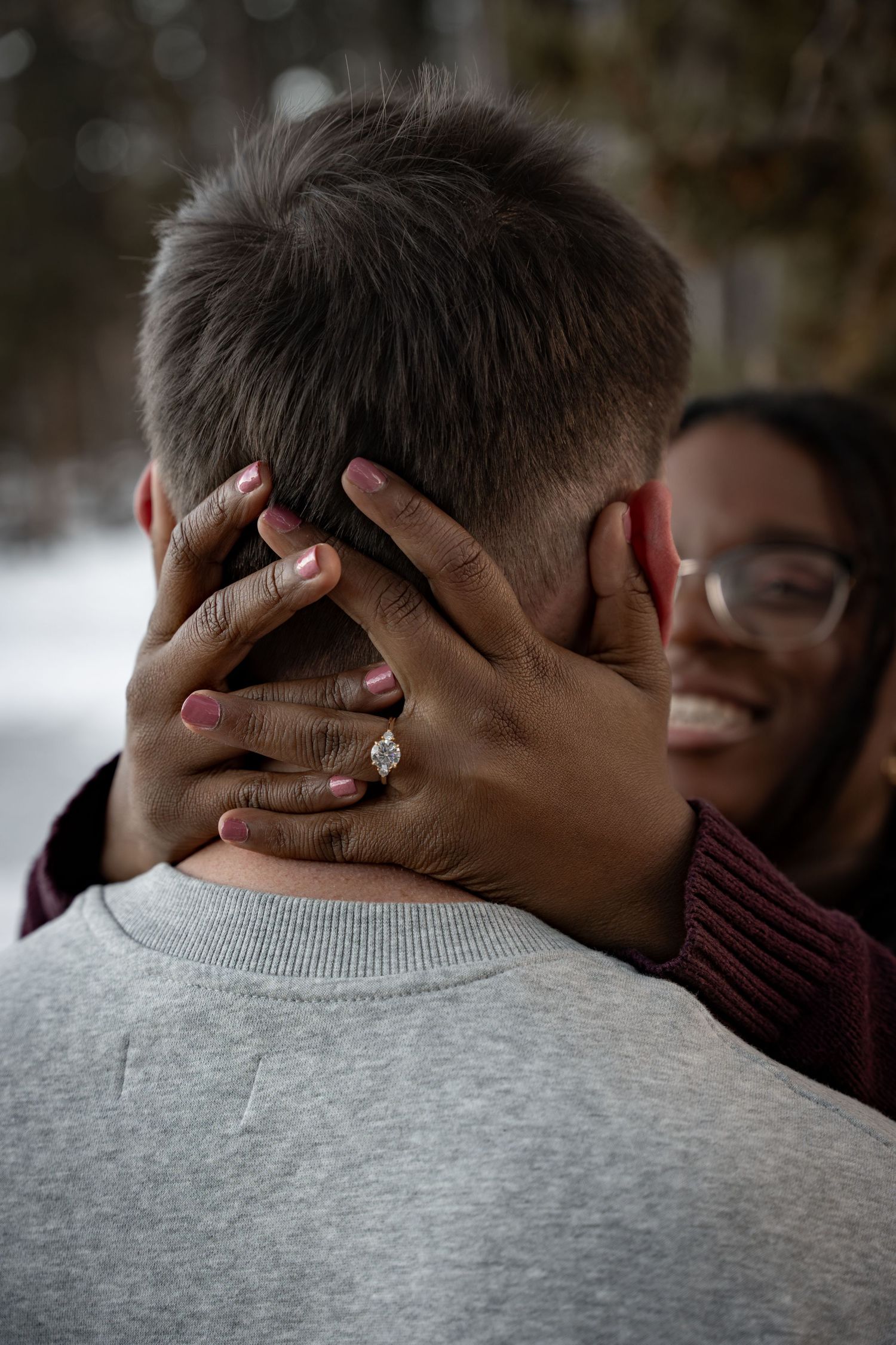 A smiling woman with glasses wearing an engagement ring embraces a man from behind in a snowy outdoor setting.