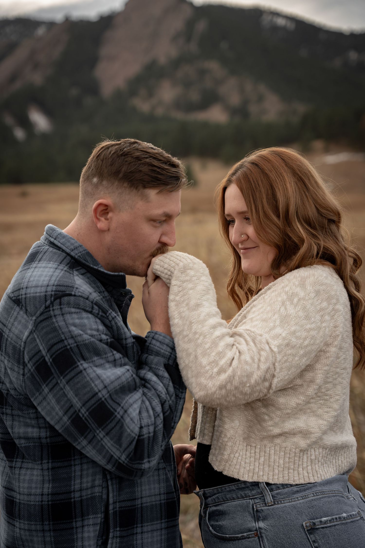 A couple shares a romantic moment outdoors with mountains in the background as he kisses her hand.