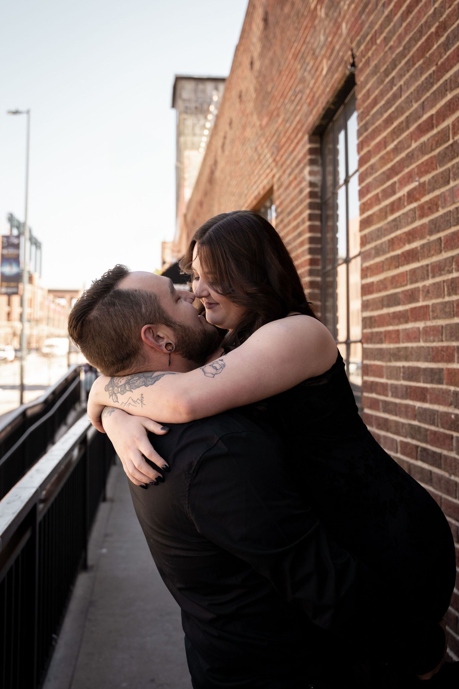 A tattooed couple in black outfits sharing a romantic embrace against a brick wall on an urban sidewalk.