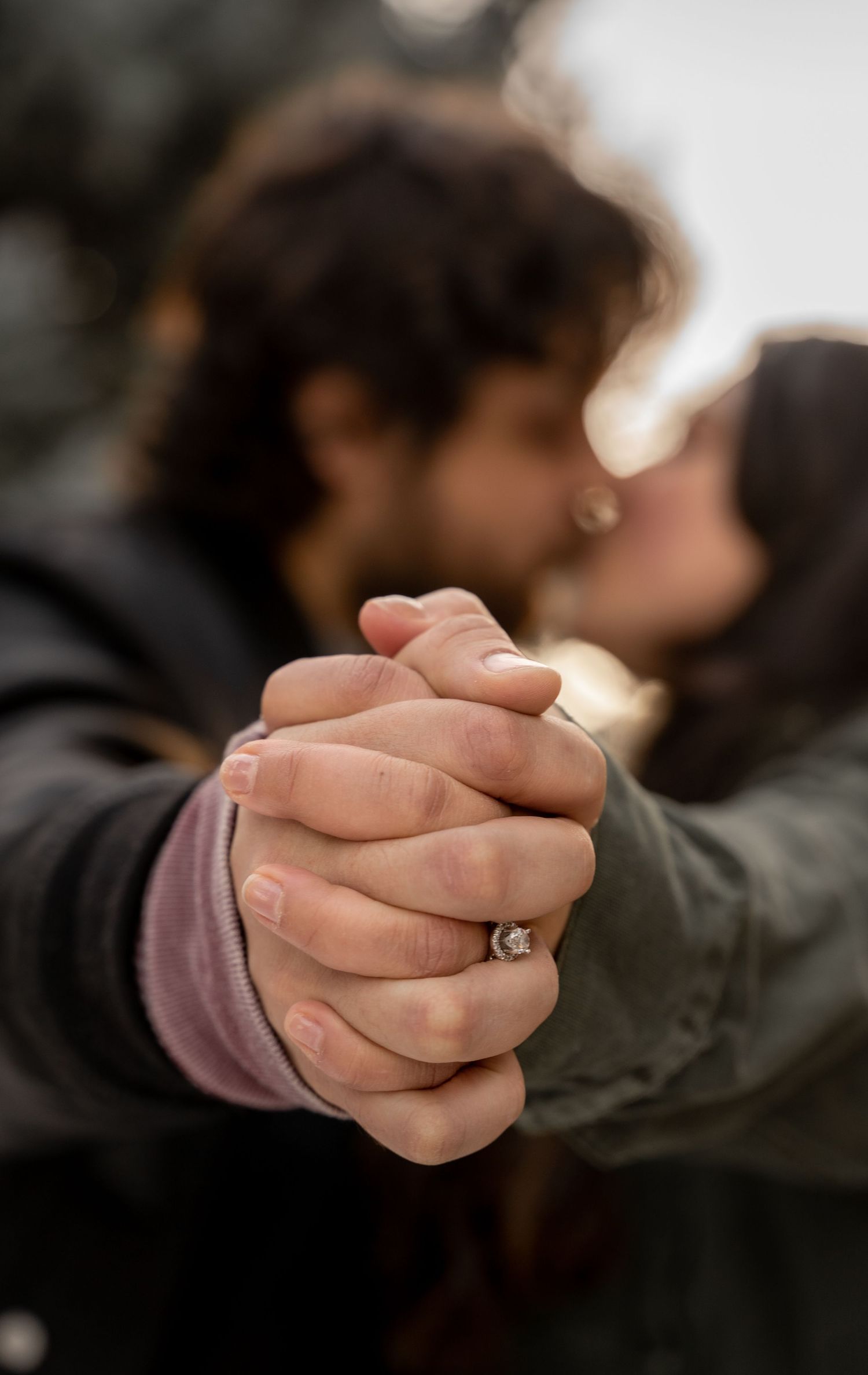 Couple kissing in background while holding hands, showcasing an engagement ring in sharp focus foreground.
