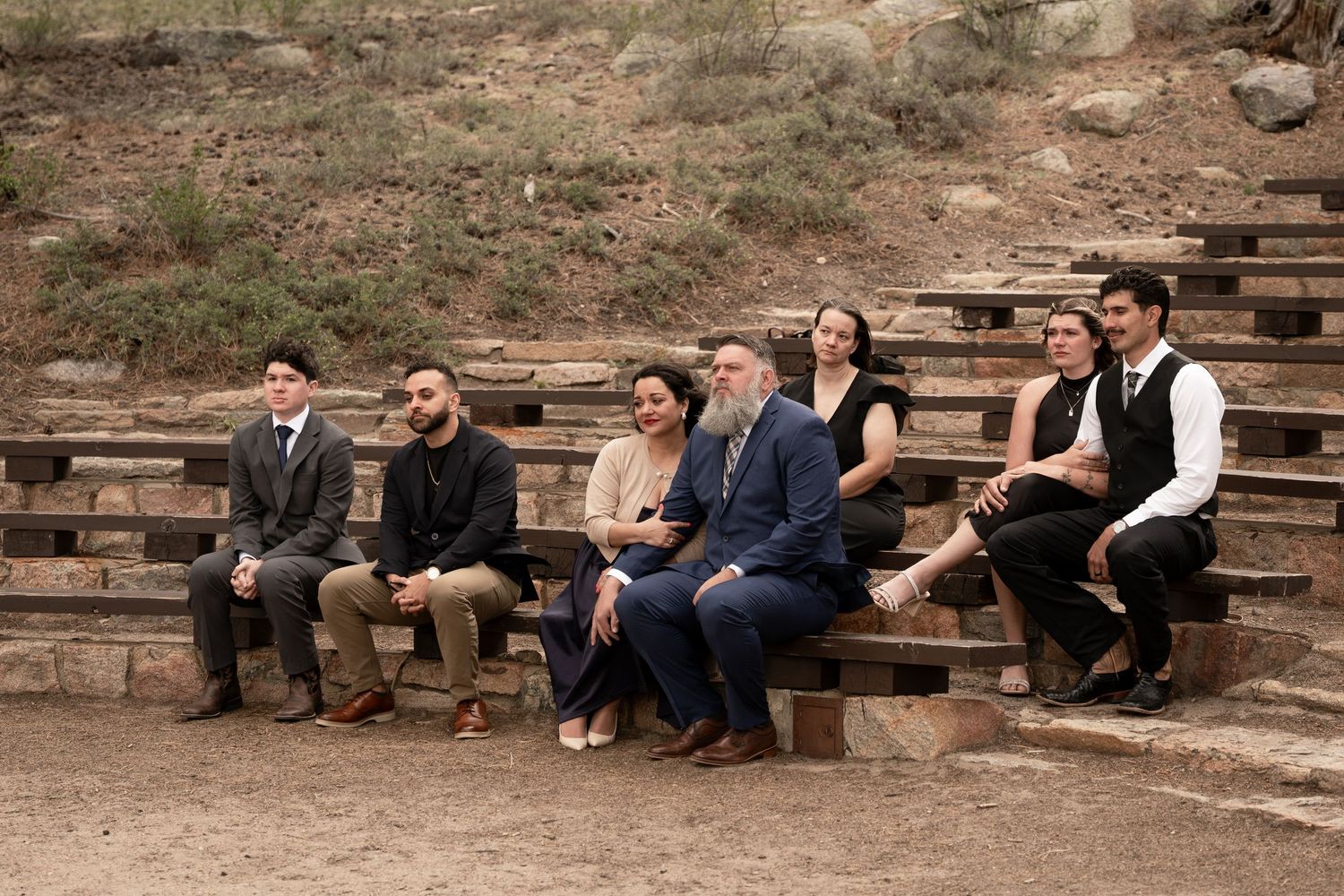 Group of seven formally dressed people sitting on rustic wooden bleachers outdoors in a desert setting.
