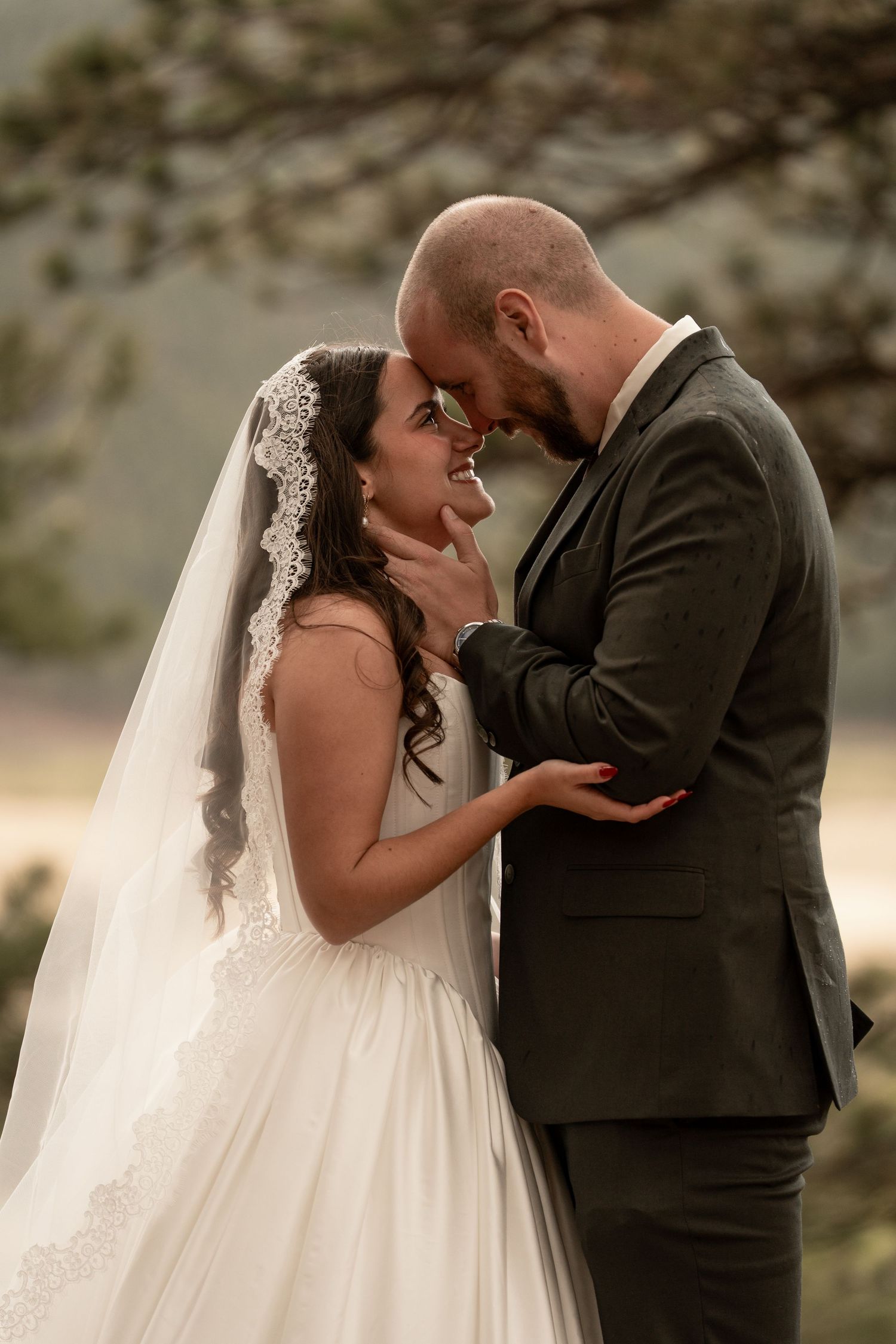 A bride and groom share a romantic forehead touch, smiling in an outdoor mountain setting on their wedding day.