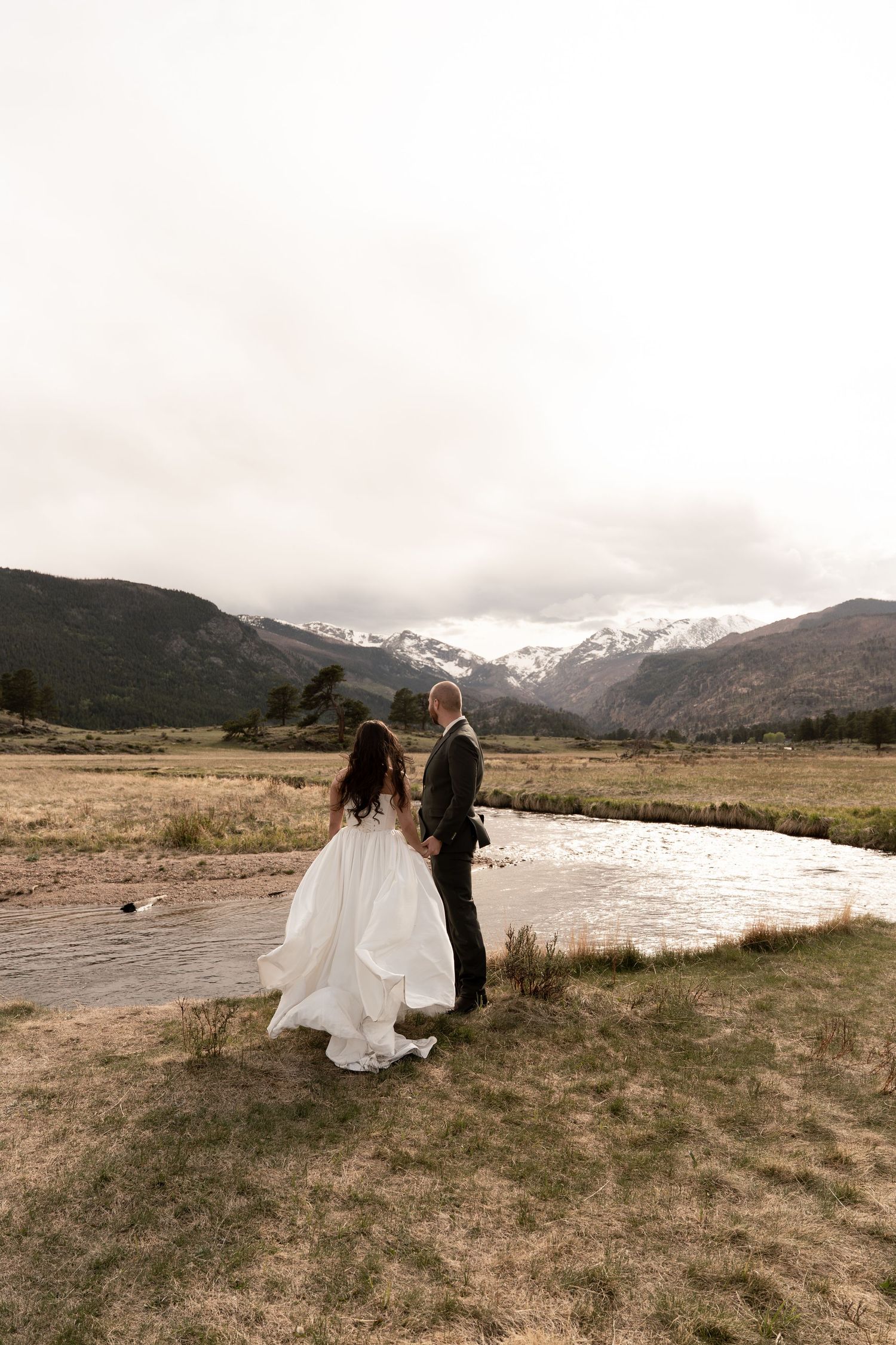 Bride and groom stand by a mountain stream with snow-capped peaks in the background during an outdoor elopement.