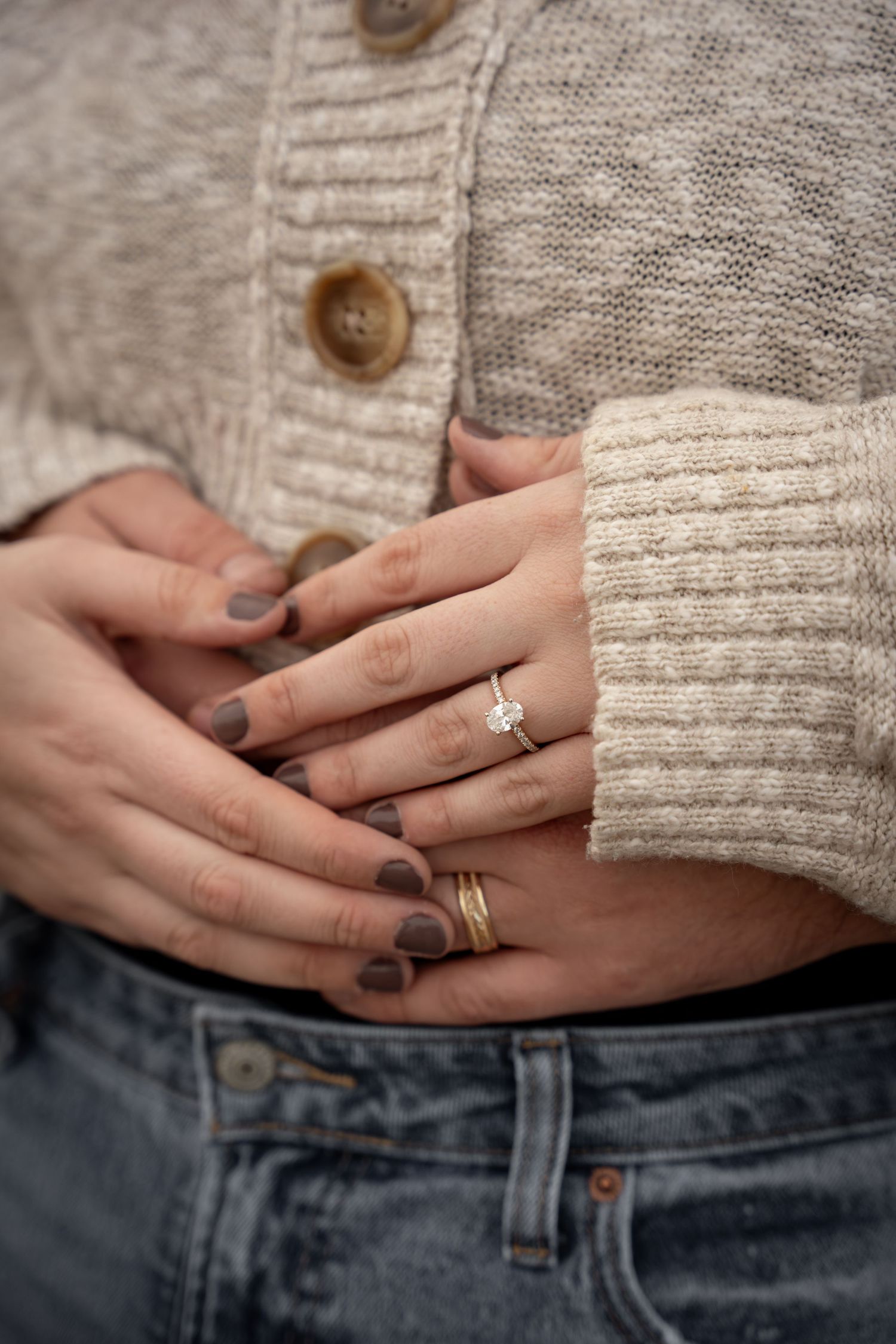 Close-up of hands with dark mauve nails wearing a diamond solitaire gold engagement ring and stacked bands.