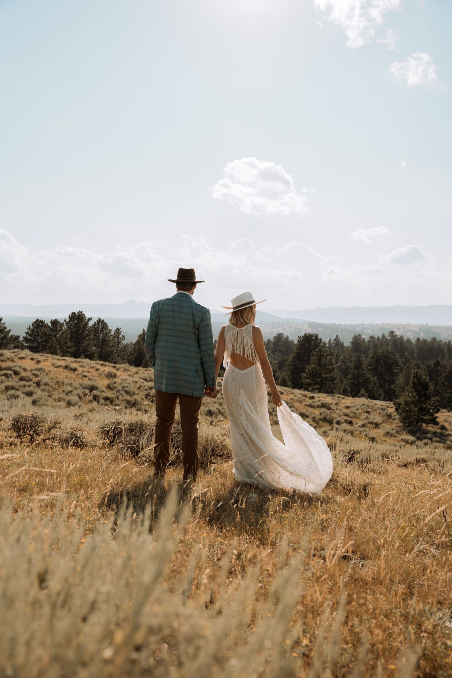 A couple walks hand in hand through a golden field with mountains and trees in the distance on a sunny day.