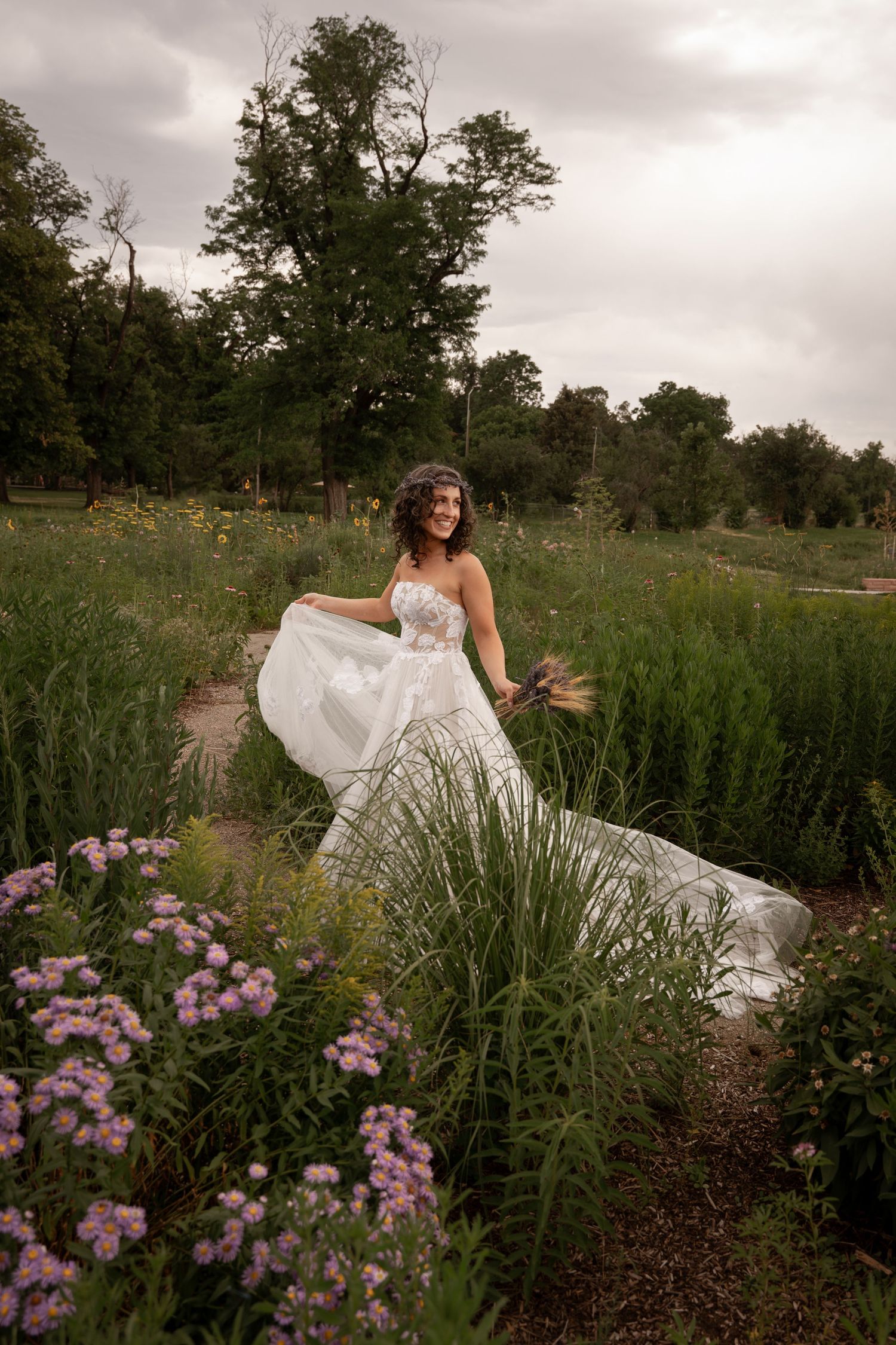 A bride in a flowing white wedding dress twirls gracefully in a wildflower garden with pink blooms and tall grasses.