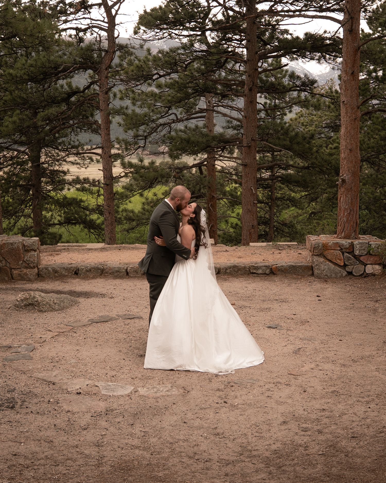 Bride and groom share a romantic kiss outdoors among tall pine trees at a rustic mountain wedding venue.